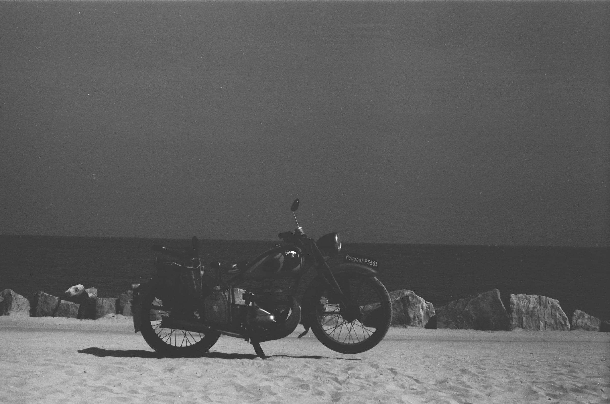 Une moto Peugeot ancienne garée sur le sable, devant les rochers de la digue.