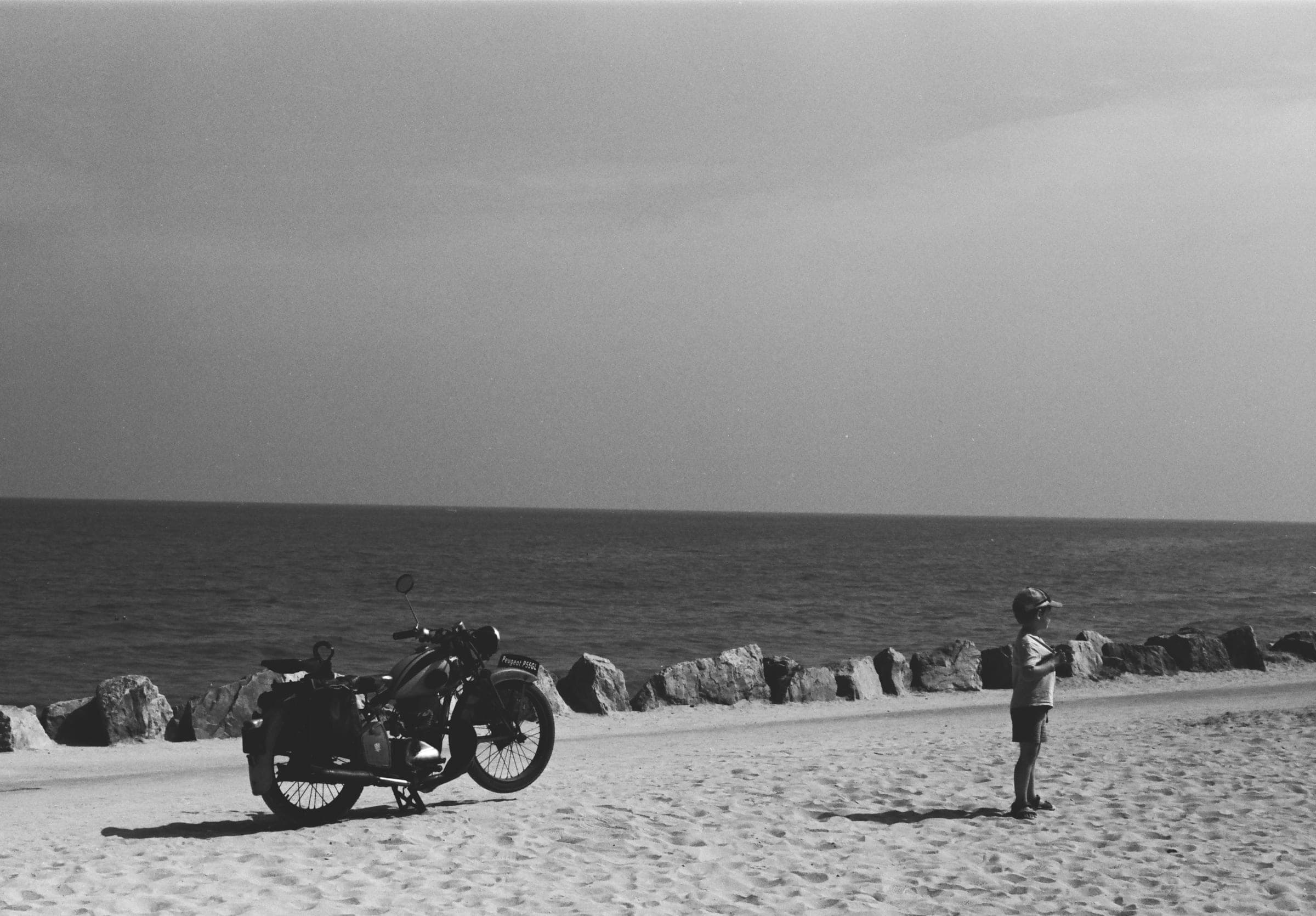 Une moto ancienne sur le sable et, quelques mètres plus loin, un petit garçon en casquette qui regarde la mer.