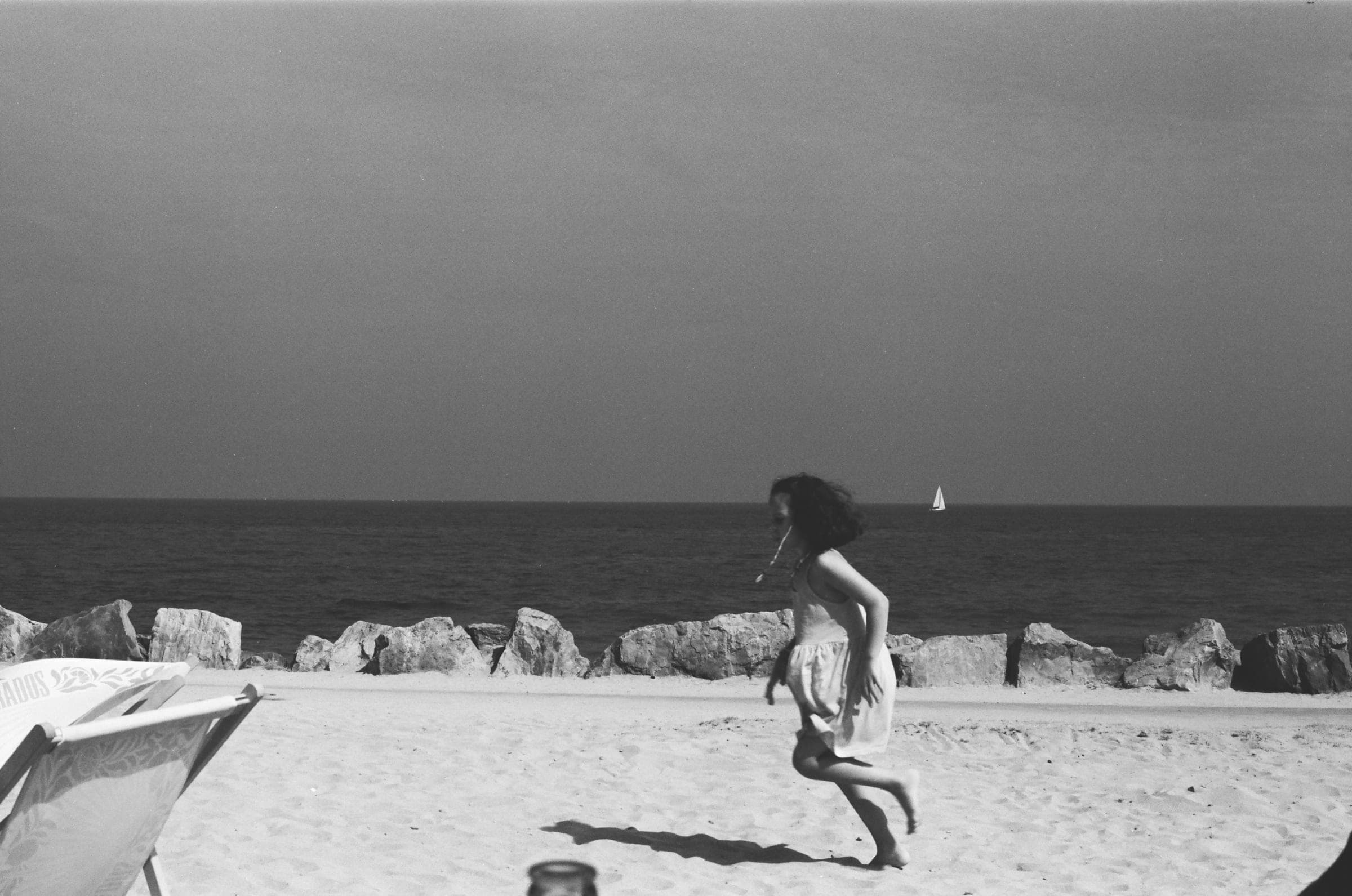 Une fille court pieds nus sur le sable, cheveux au vent, un voilier au loin.