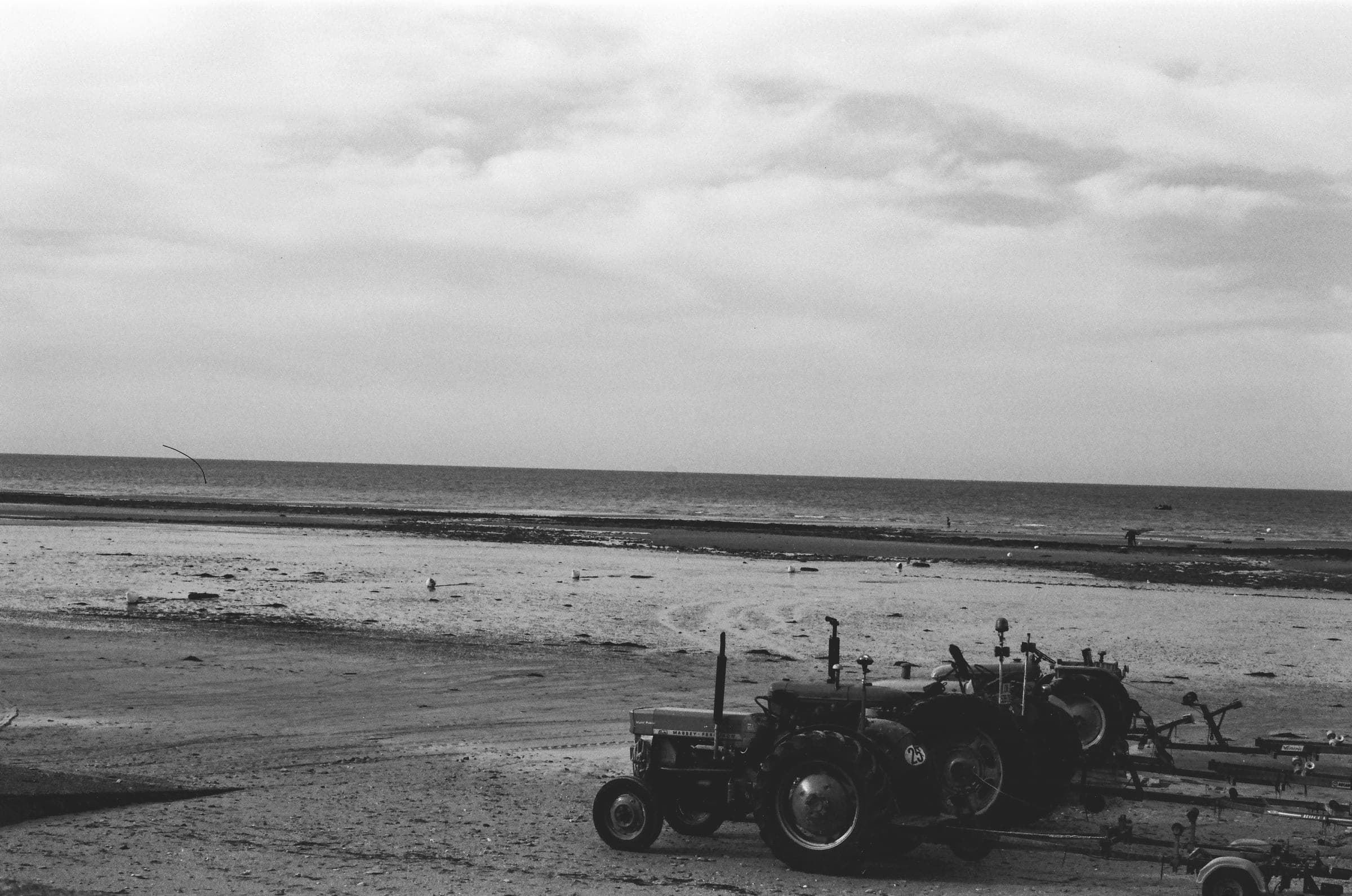Des tracteurs alignés sur leurs remorques au bord de l'estran, sous un ciel chargé.