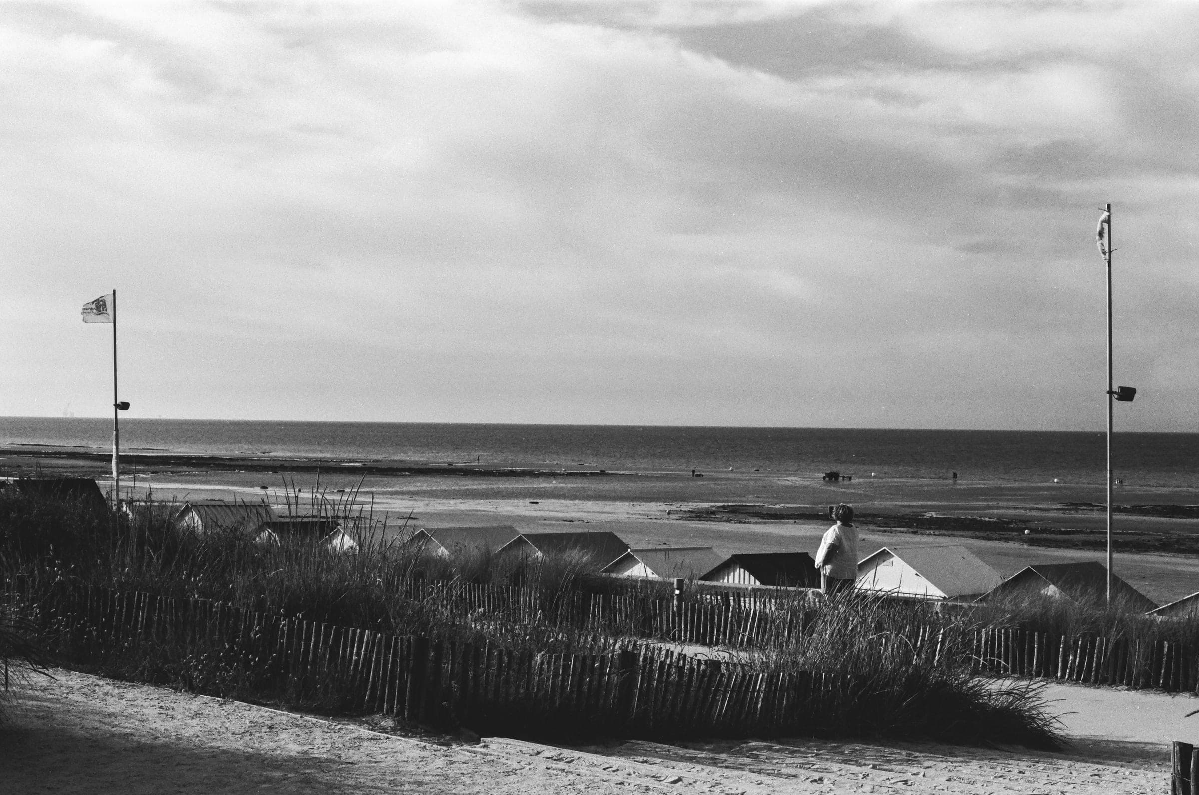 Une silhouette de dos au-dessus des toits des cabines, derrière les ganivelles et les oyats, la mer basse au loin.