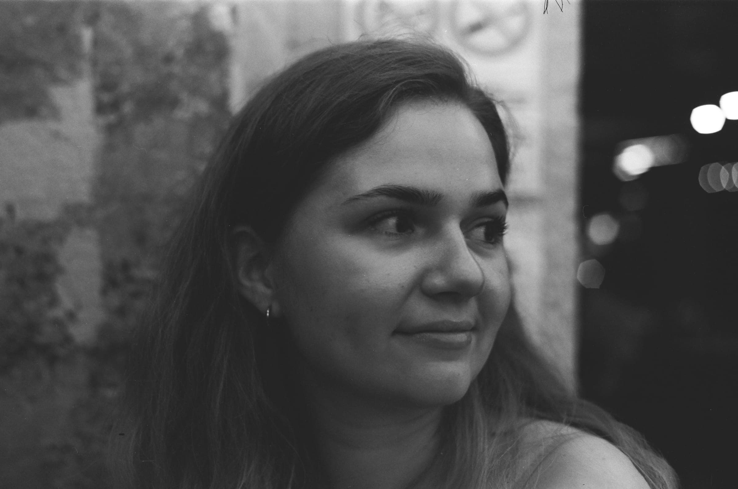 Portrait d'une femme le soir, le regard de côté et un léger sourire, des lumières floues derrière elle.