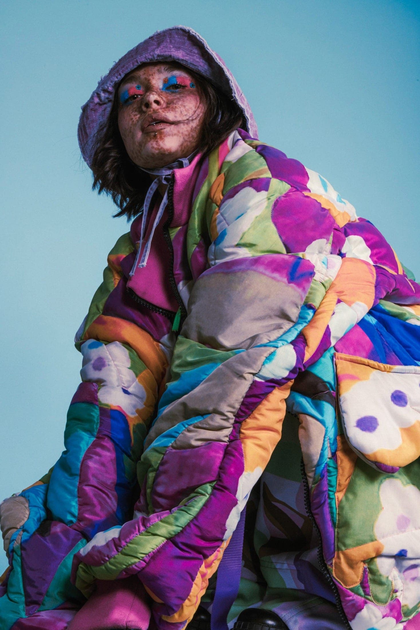 Bob lilas et doudoune matelassée à motifs floraux multicolores, fard bleu et rouge sur les paupières, fond de studio bleu.