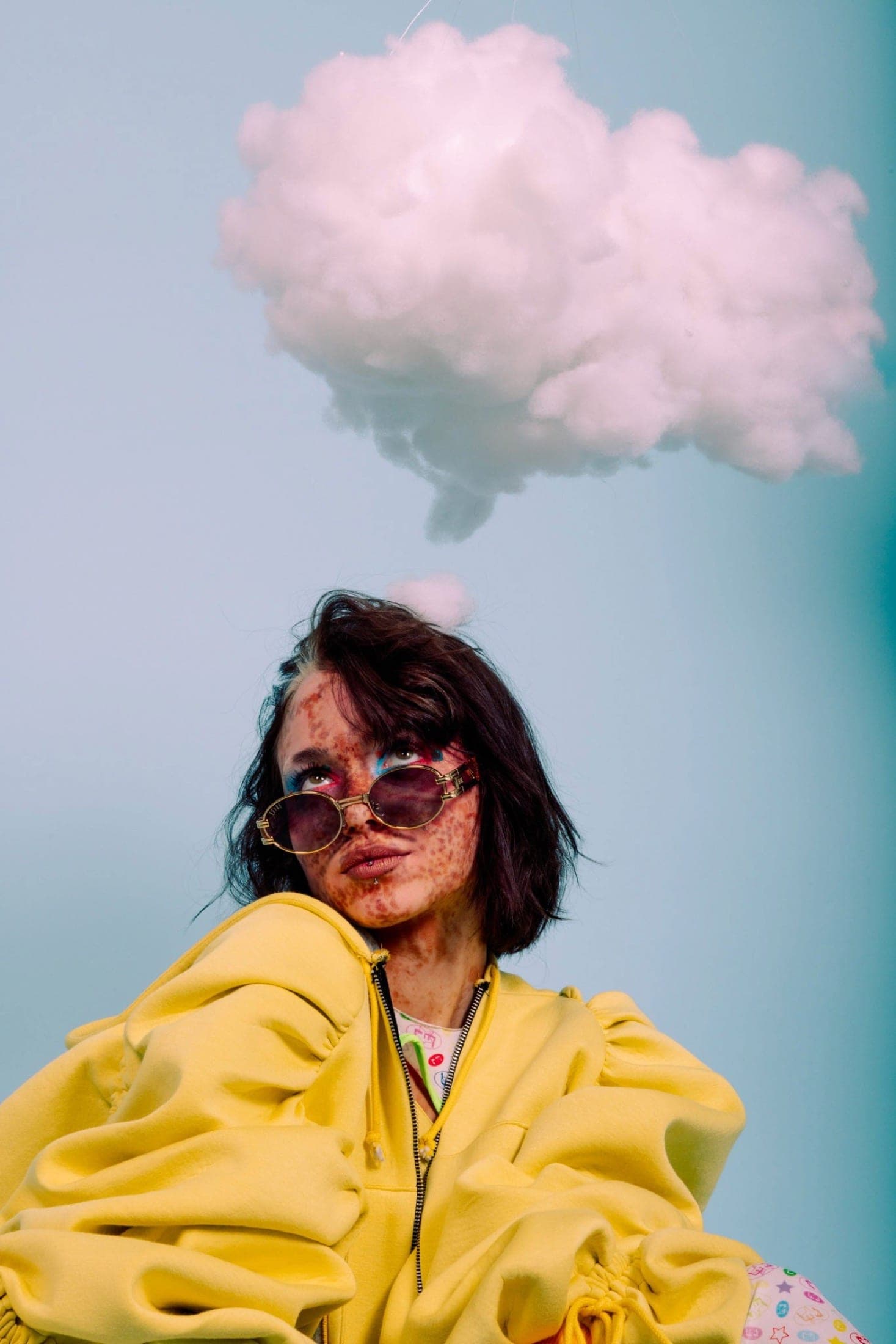 Un nuage de ouate suspendu au-dessus de la tête, sweat jaune à capuche et lunettes ovales fumées, le regard levé vers le nuage.