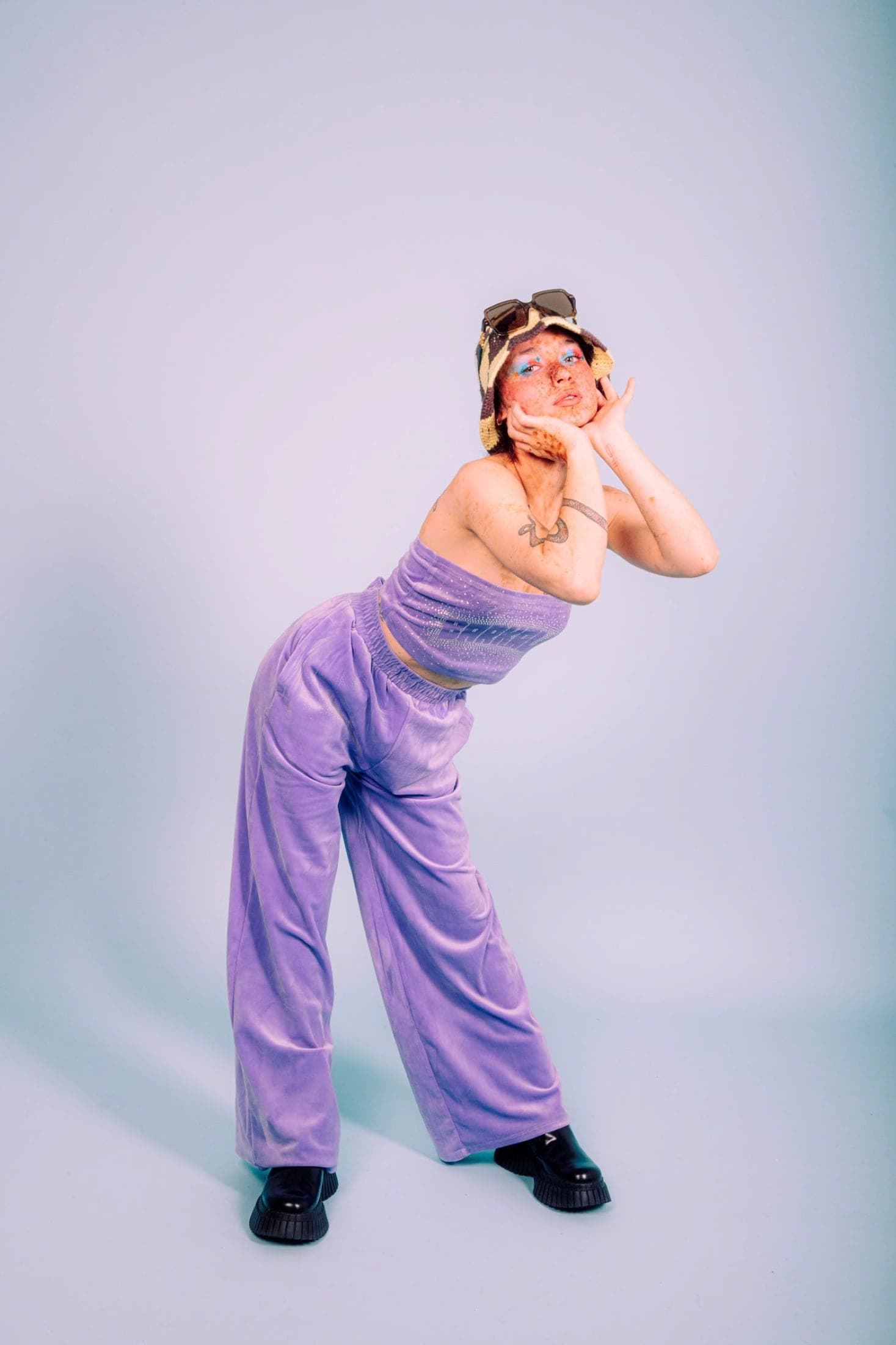 Plan en pied sur fond bleu : ensemble en velours lilas, bob crocheté, bottines noires à grosse semelle et tatouage de serpent sur le bras.