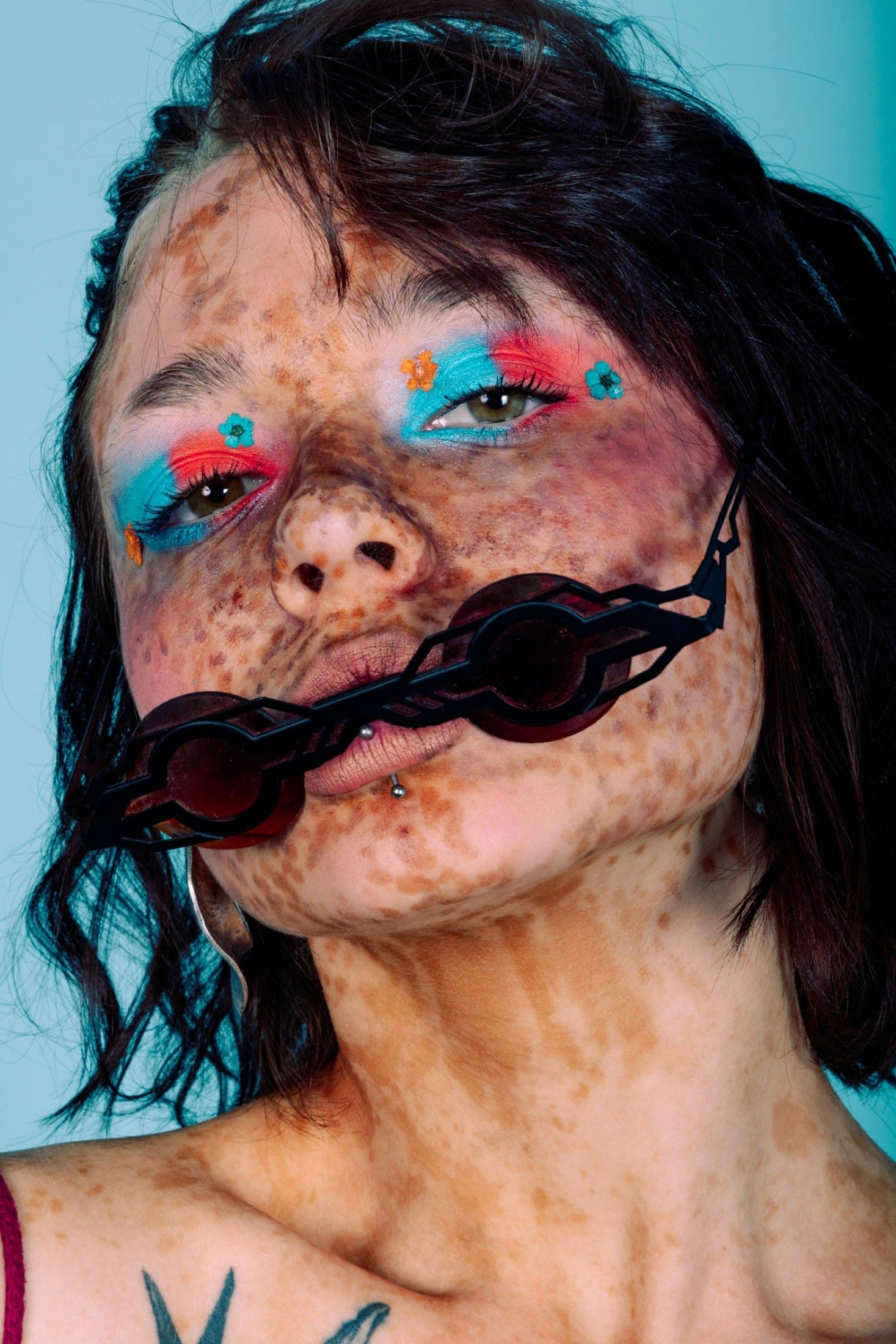 Gros plan sur un visage aux taches de rousseur denses, fard bleu et rouge parsemé de petites fleurs, lunettes noires serrées entre les dents.