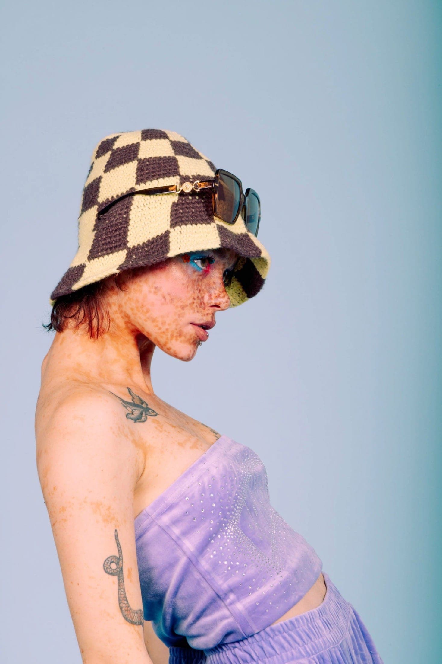 De profil, bob crocheté à damier crème et brun surmonté de lunettes, bandeau de velours lilas, tatouages d'hirondelle et de serpent.