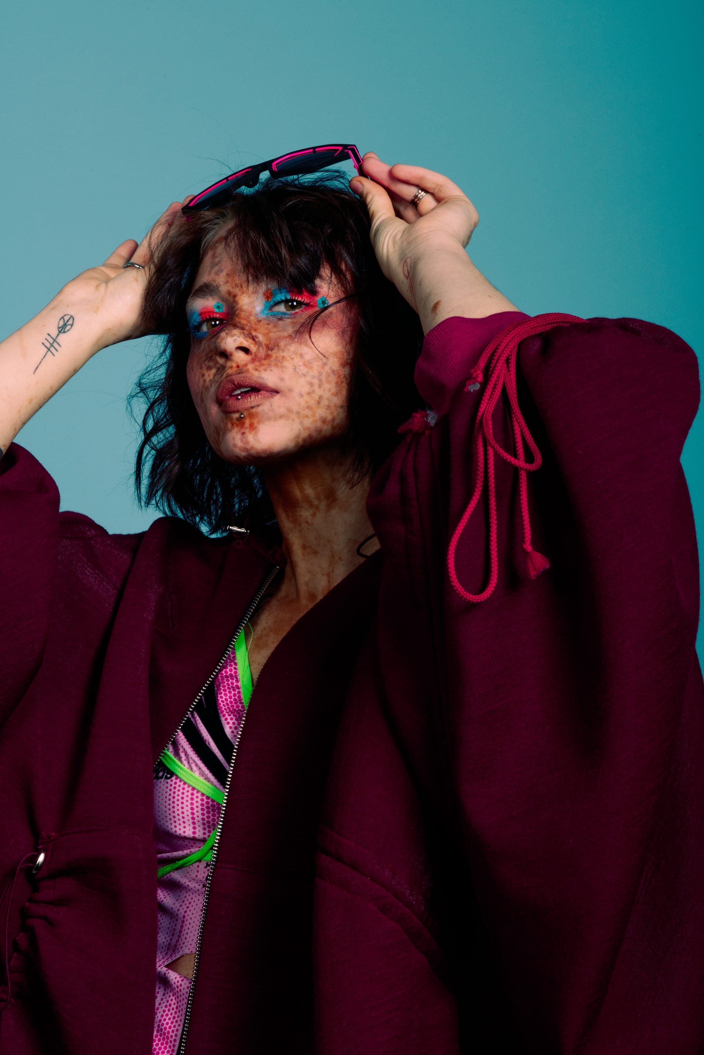 Mains dans les cheveux, lunettes roses remontées sur le front, fard bleu et rouge, veste bordeaux à cordons roses.