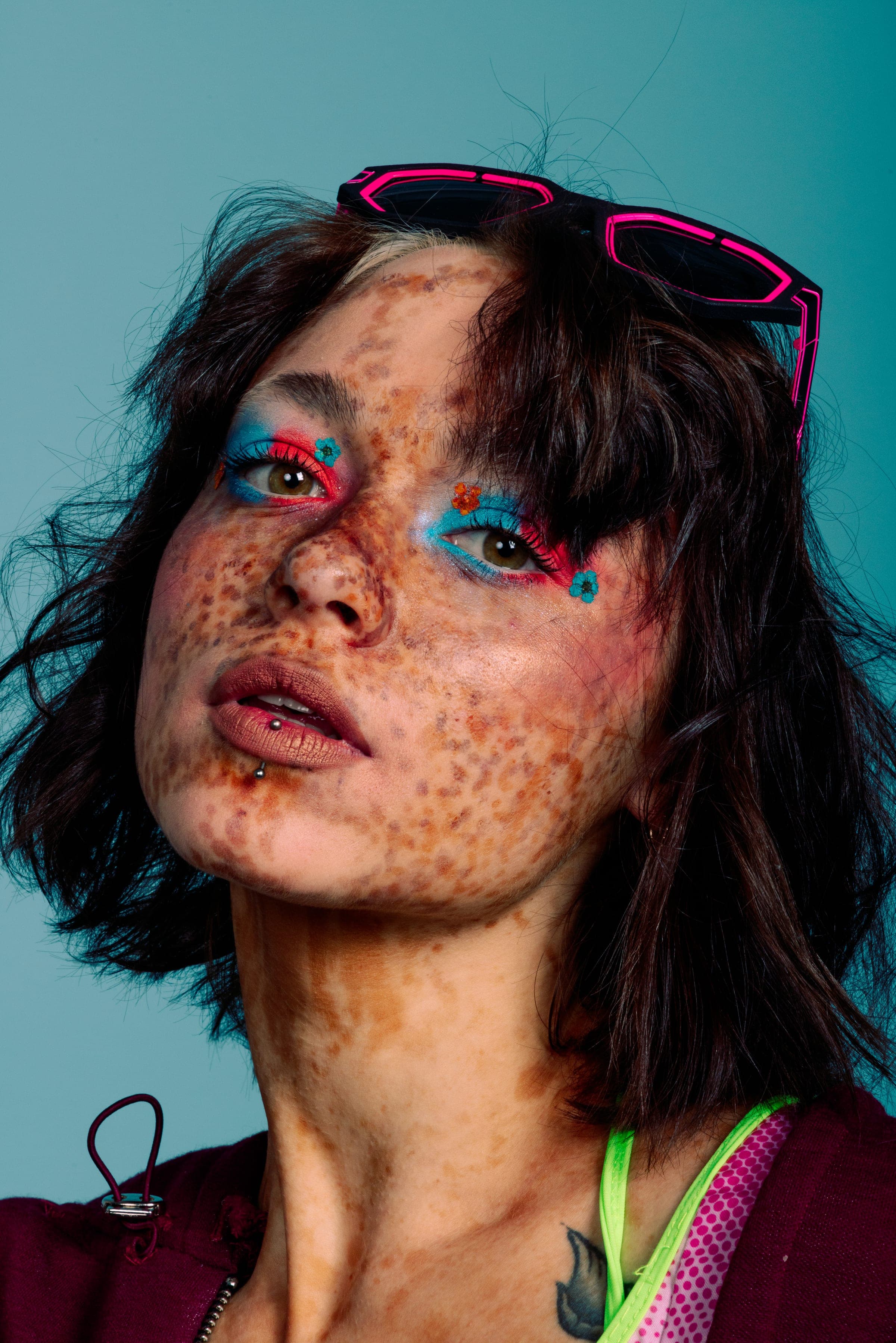 Visage levé en gros plan, taches de rousseur jusqu'au cou, fard bleu et rouge semé de petites fleurs, lunettes roses posées dans les cheveux.