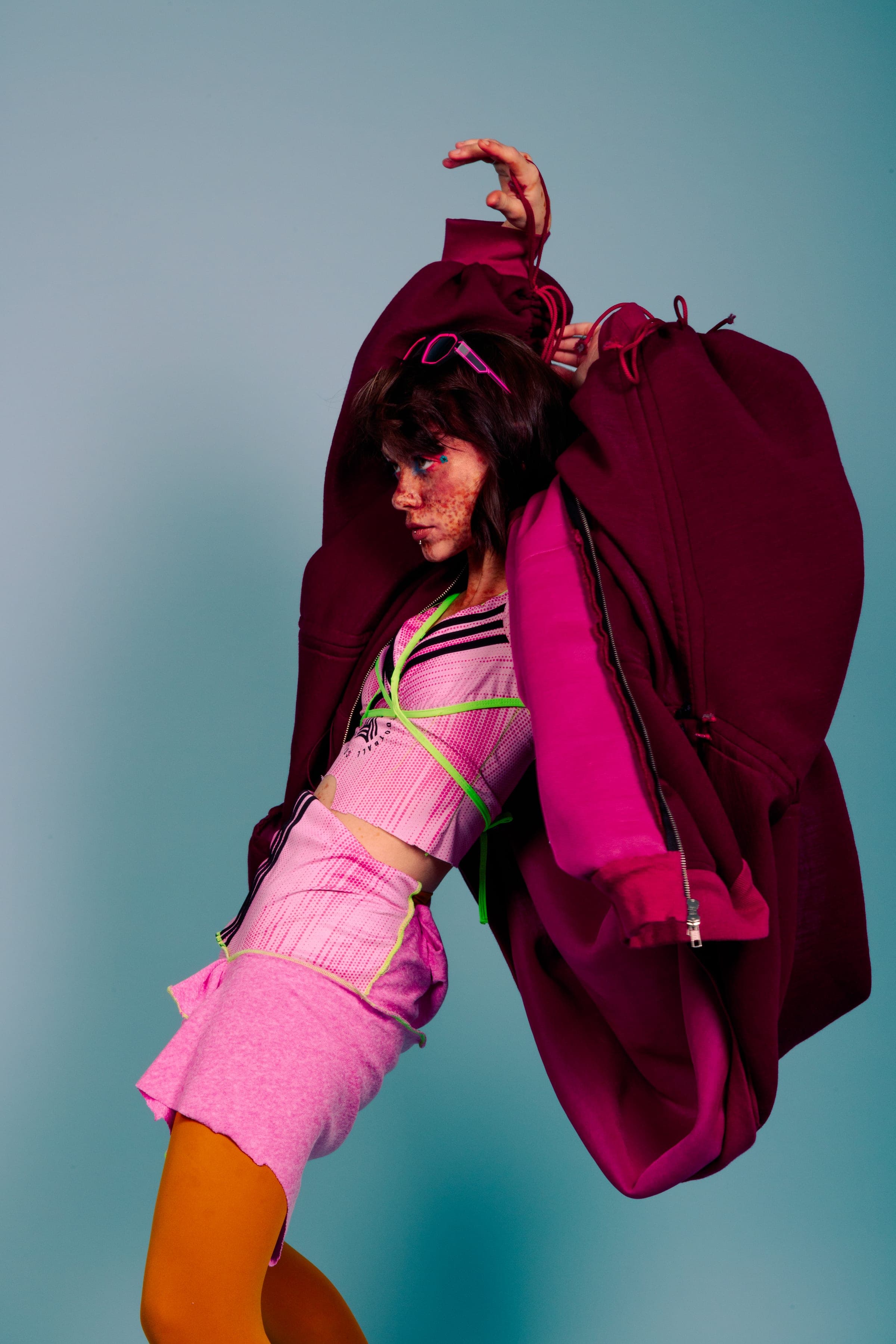 Bras levés dans un sweat bordeaux trop grand qui retombe comme une cape, haut rose rayé de noir et de vert fluo, collants orange.