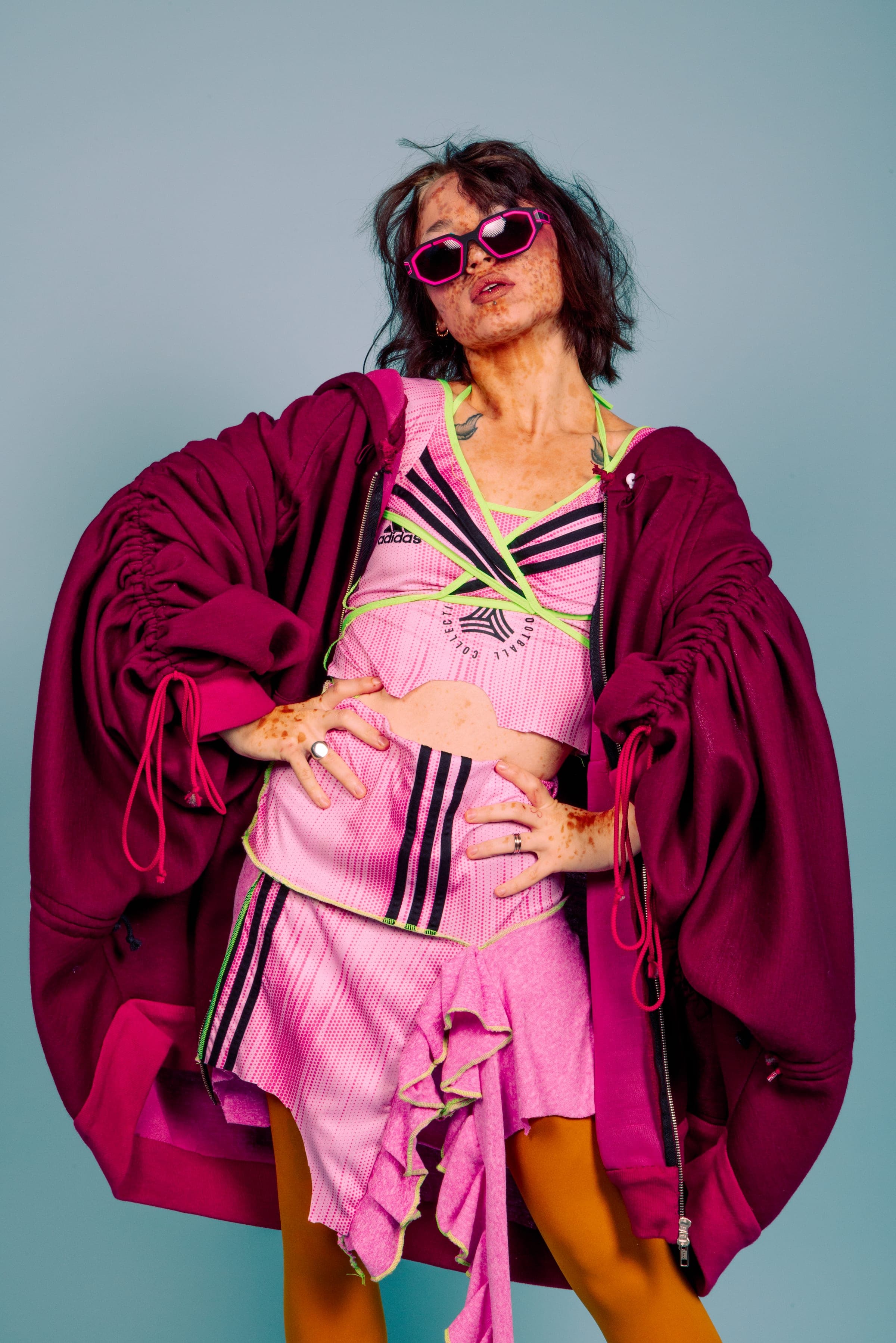 Mains sur les hanches et lunettes roses, ensemble rose à rayures noires bordé de vert fluo, veste bordeaux froncée, collants orange.
