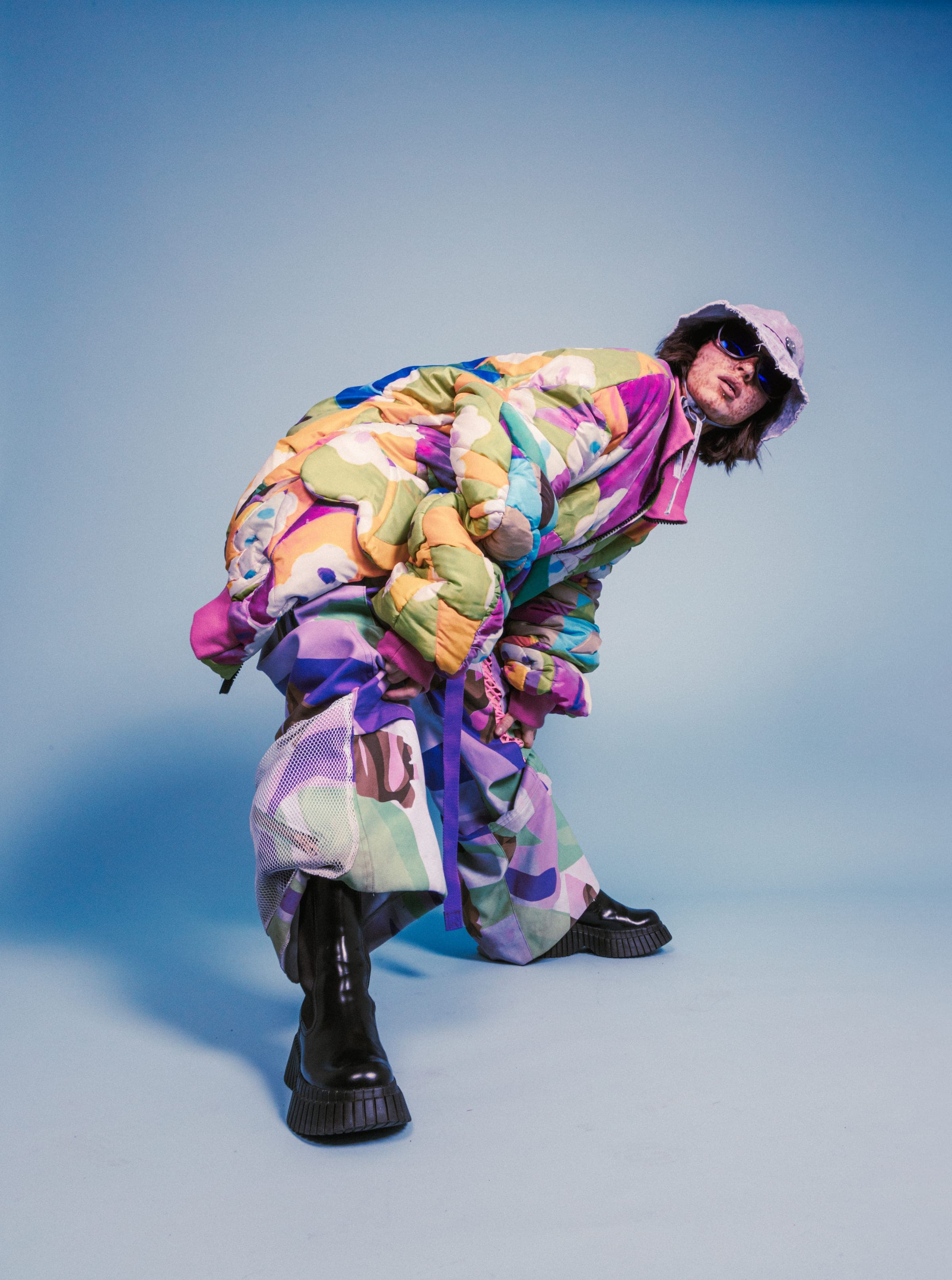 Accroupie de profil dans une doudoune à fleurs pastel, bob lilas, lunettes noires et grosses bottes vernies.