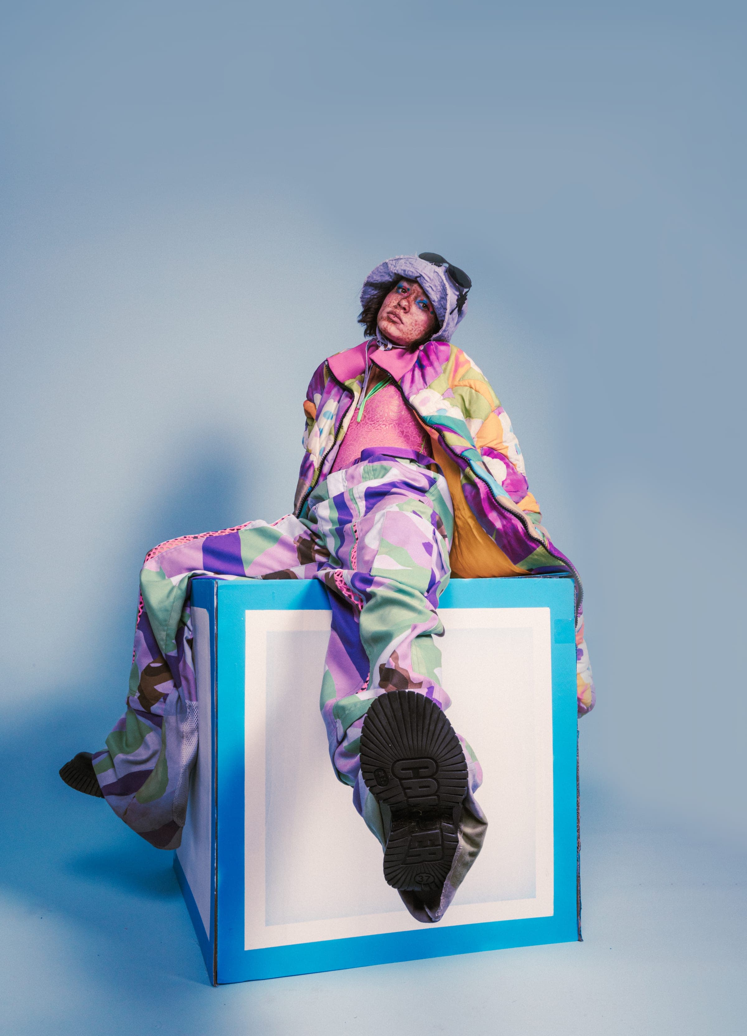 Assise sur une boîte en carton bleu et blanc, une semelle crantée tendue vers l'objectif, doudoune et pantalon à fleurs.