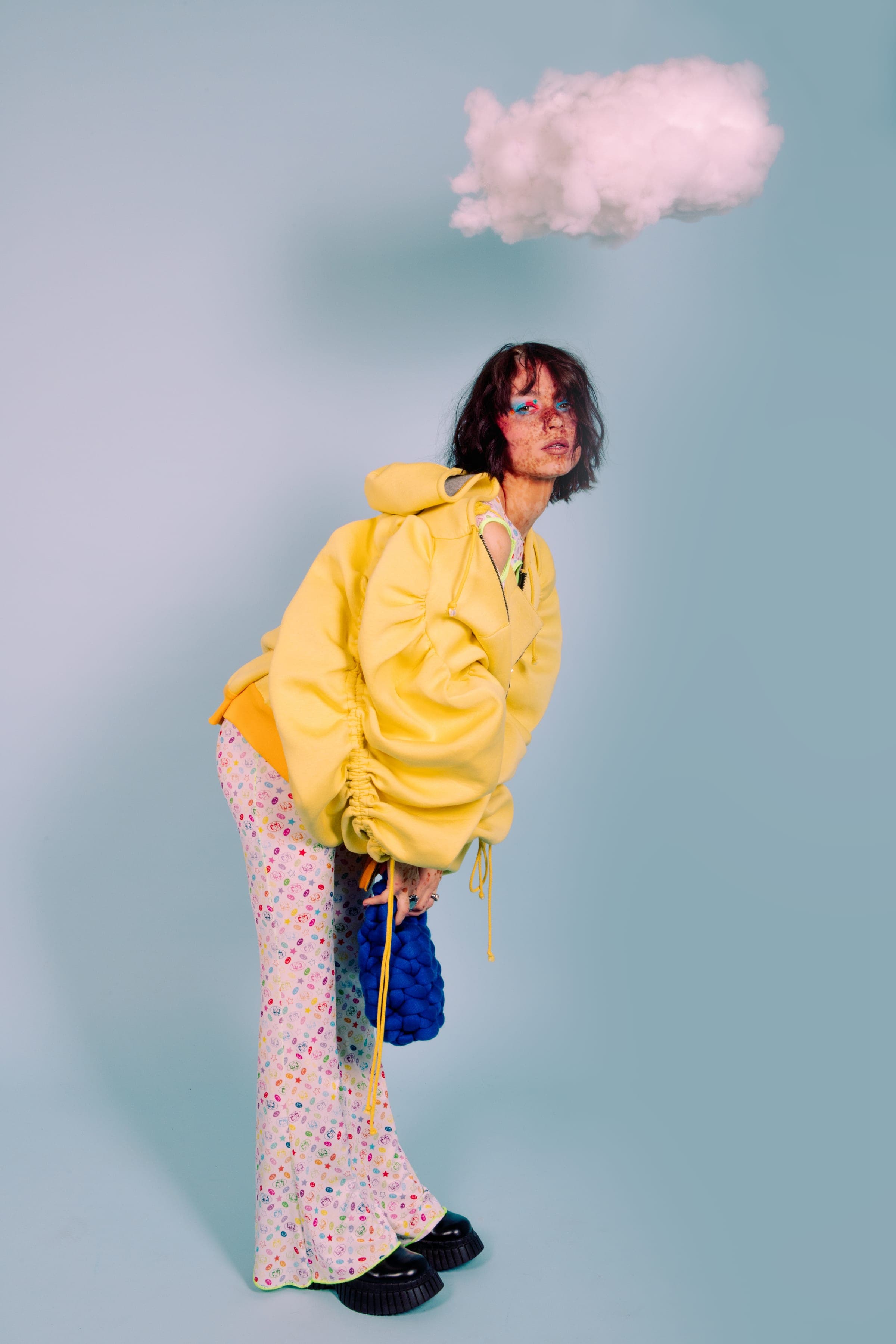 Penchée en avant sous un nuage de ouate, une mèche dressée vers lui, veste jaune froncée, sac tricoté bleu et pantalon évasé à pois.