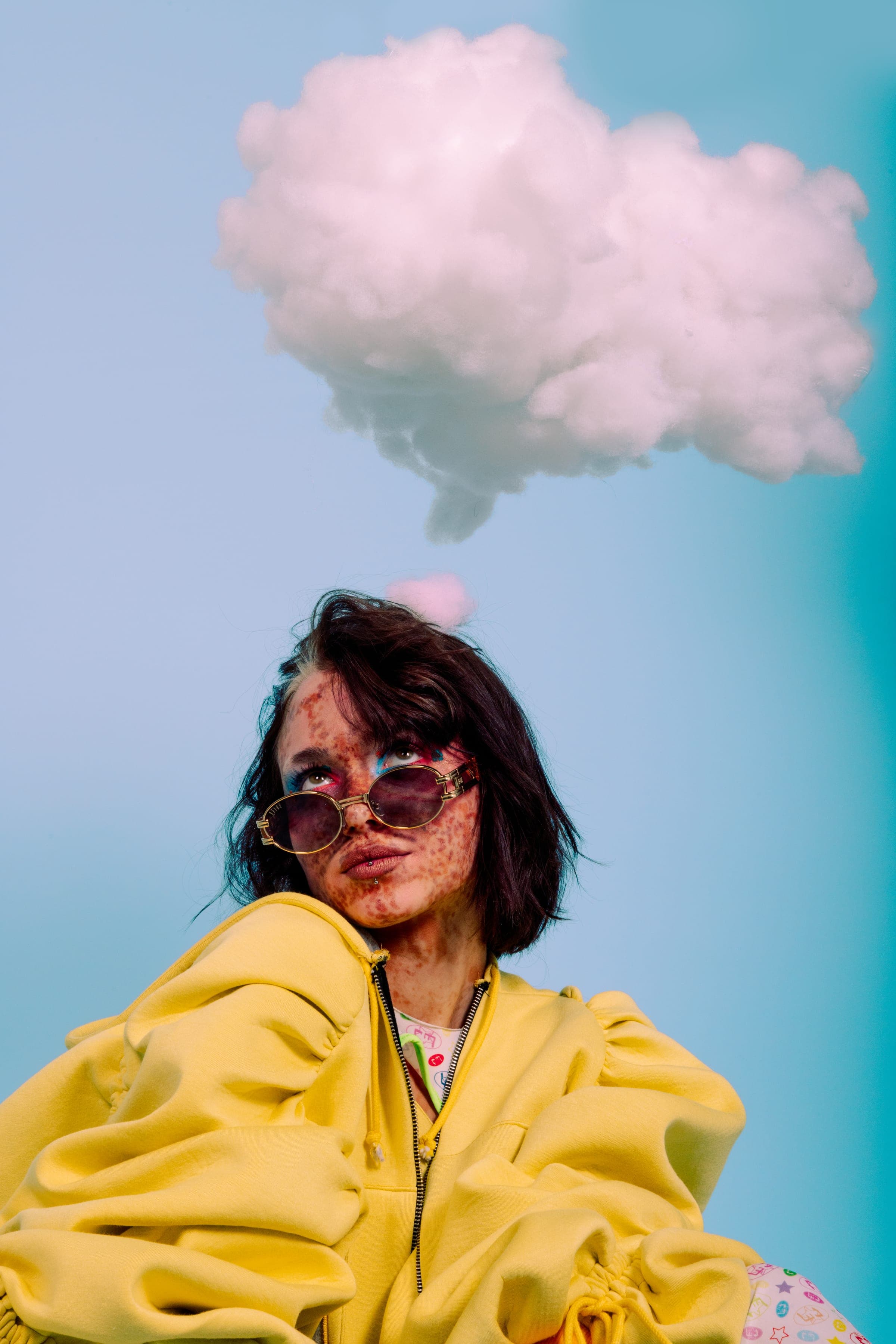 Les yeux levés vers un gros nuage blanc, lunettes rondes dorées et bouche en moue, col d'une veste jaune volumineuse.