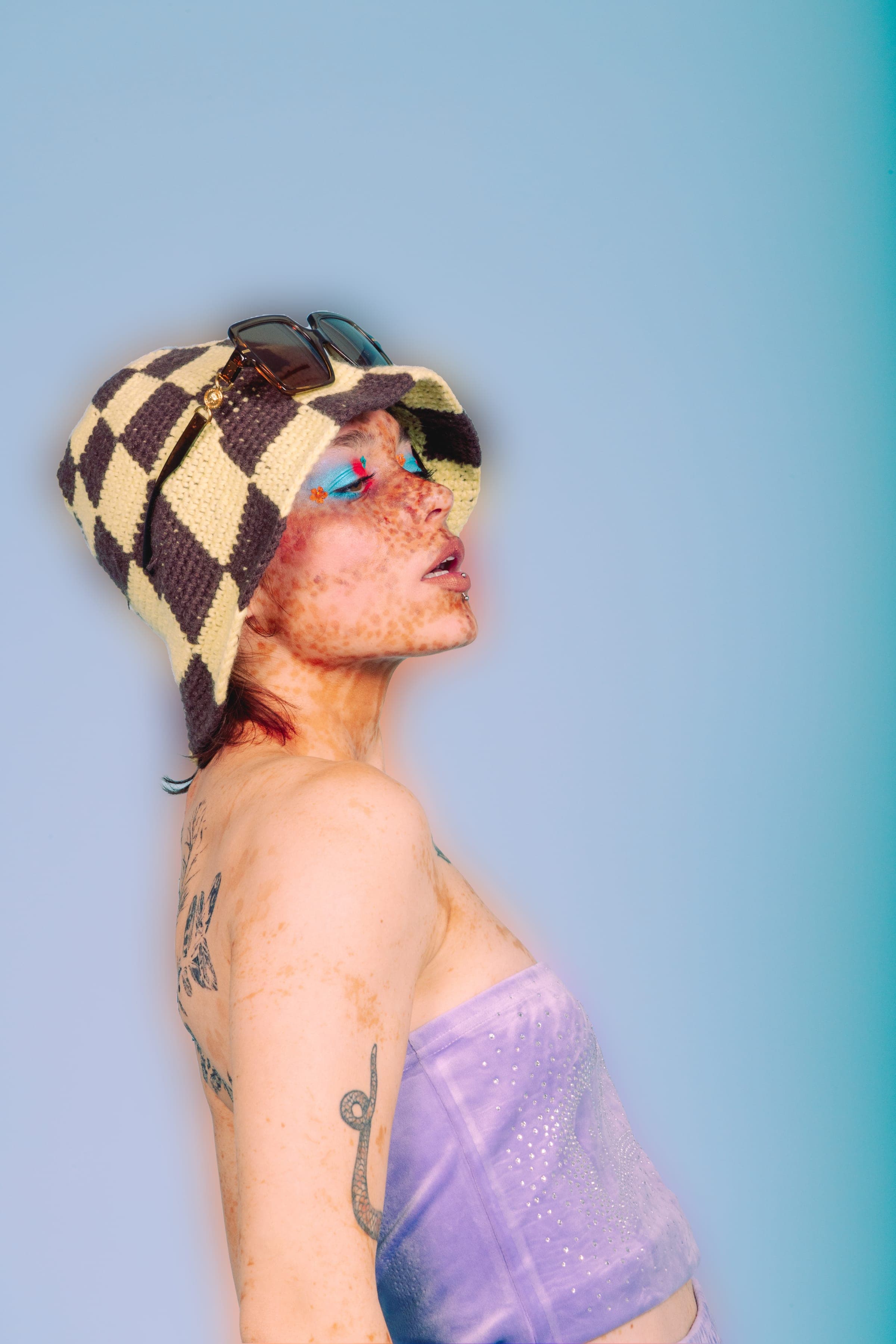 De profil, les yeux mi-clos, bob à damier surmonté de lunettes, bandeau de velours lilas, serpent tatoué le long du bras.