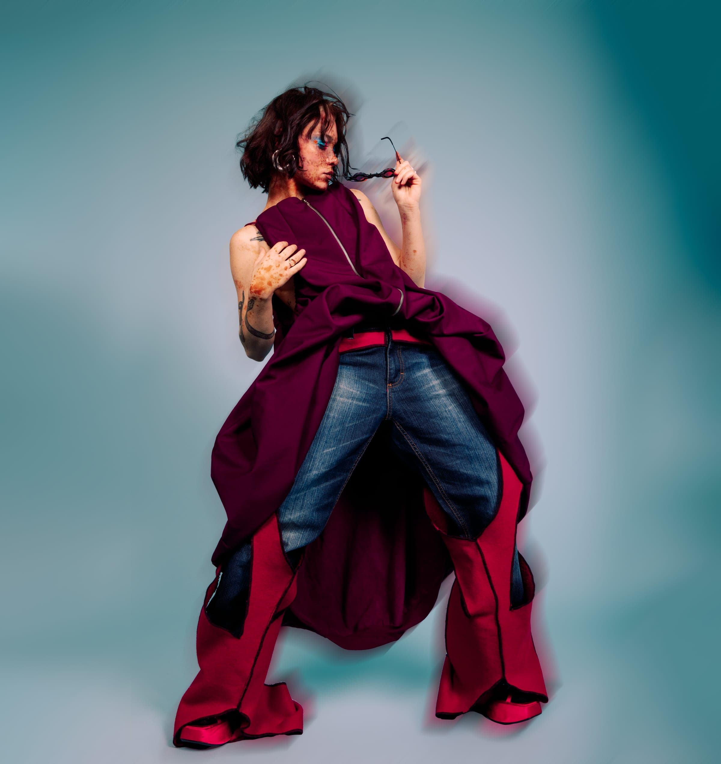 Robe bordeaux retroussée sur un jean, jambières rouges évasées, une branche de lunettes pincée entre deux doigts.