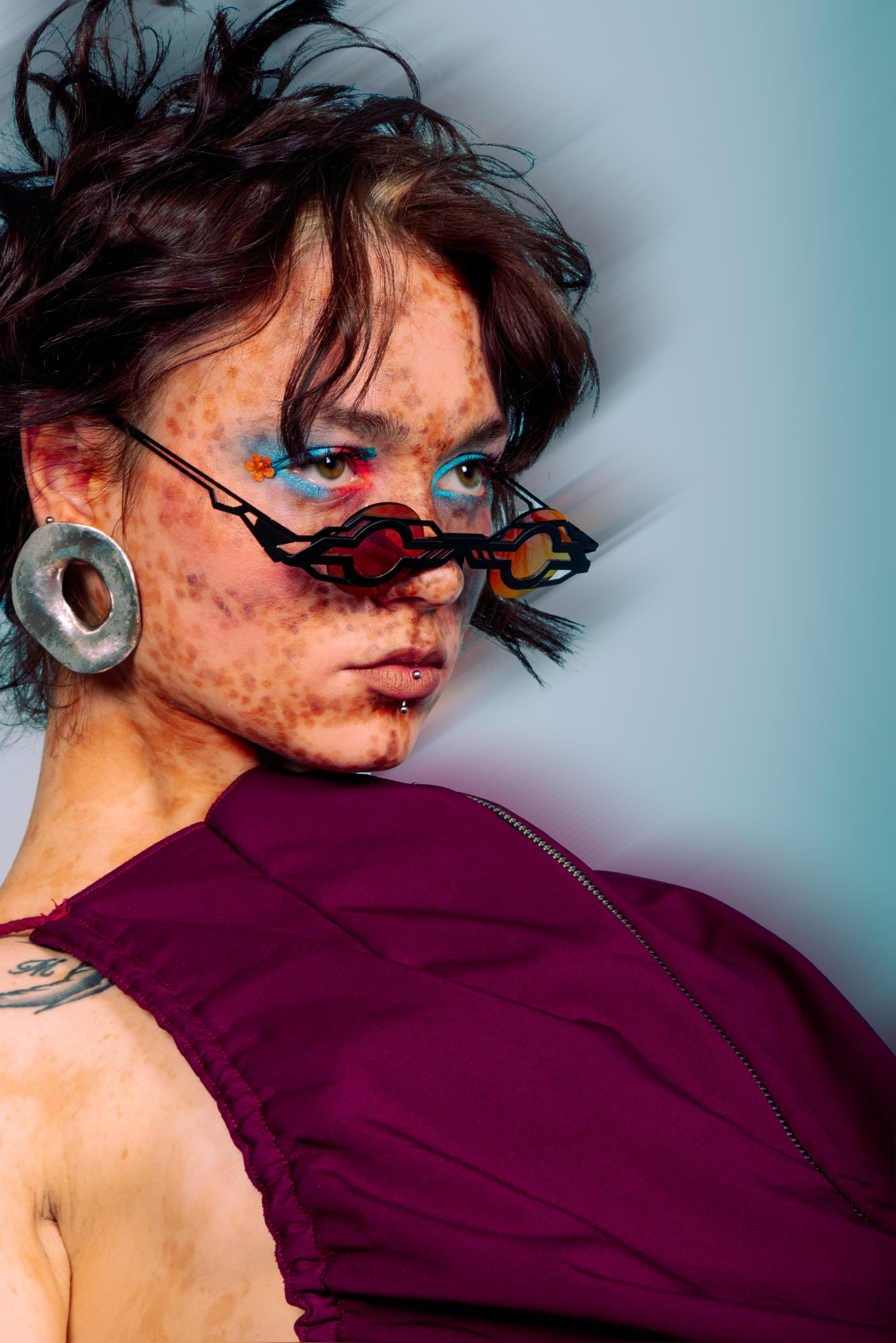 Regard en coin par-dessus des lunettes noires ajourées posées bas sur le nez, grand anneau d'argent à l'oreille, robe bordeaux.