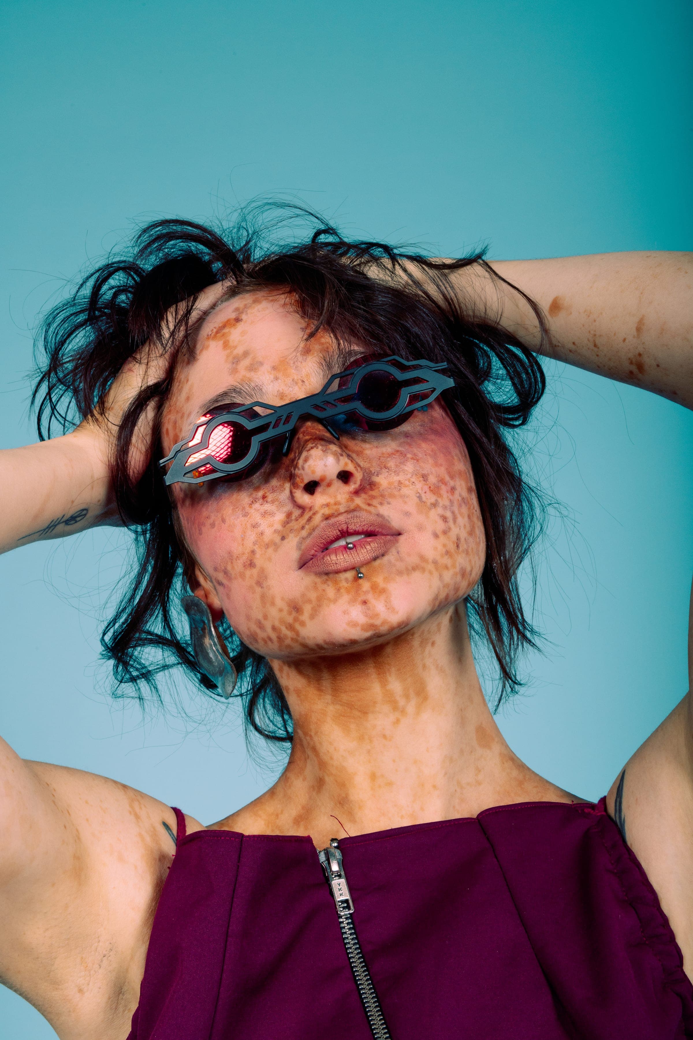 Mains dans les cheveux en bataille, lunettes noires découpées aux verres rouges, taches de rousseur sur le cou et les bras, robe bordeaux zippée.