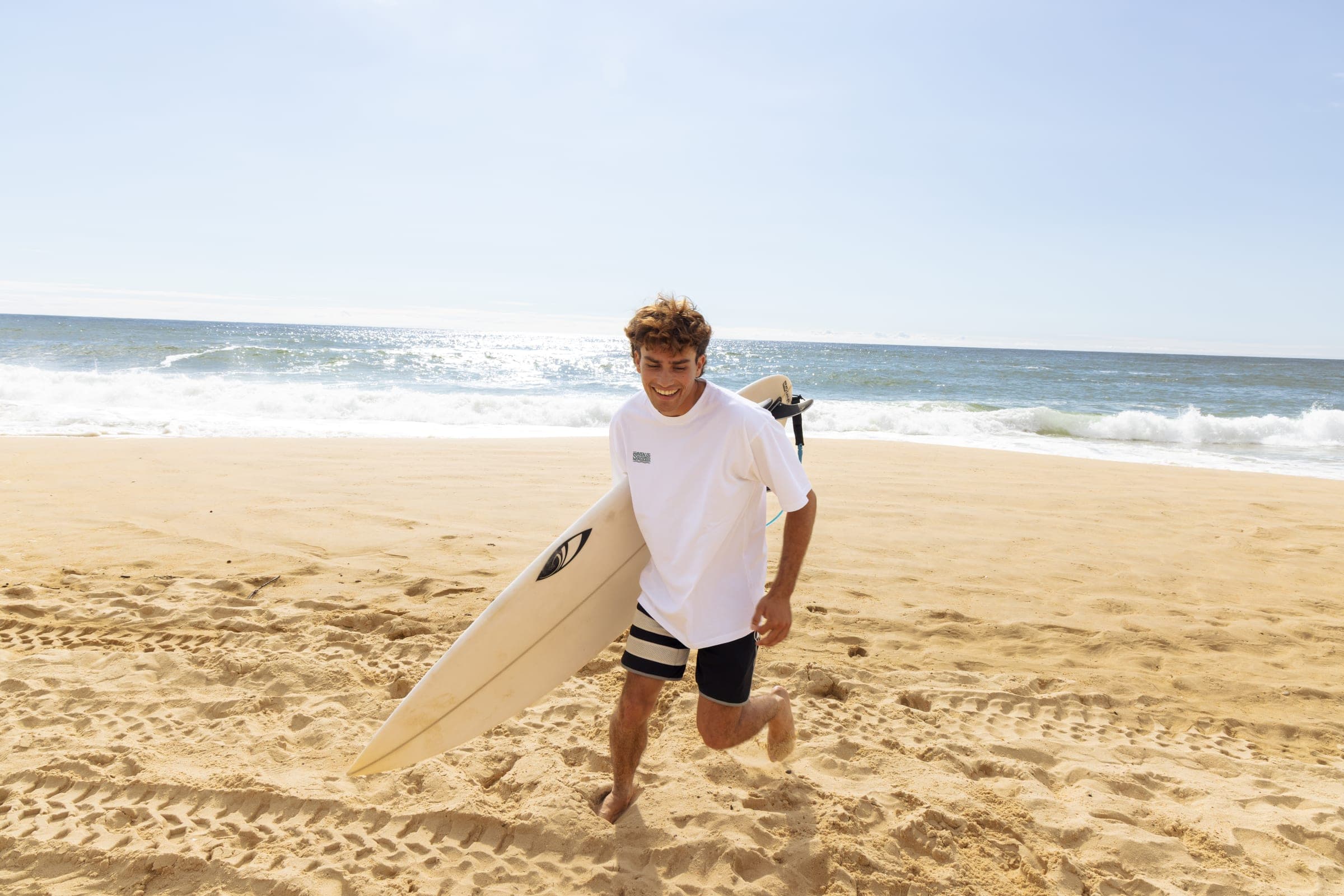 Un surfeur remonte la plage, planche sous le bras, en t-shirt blanc Uma Rosie.