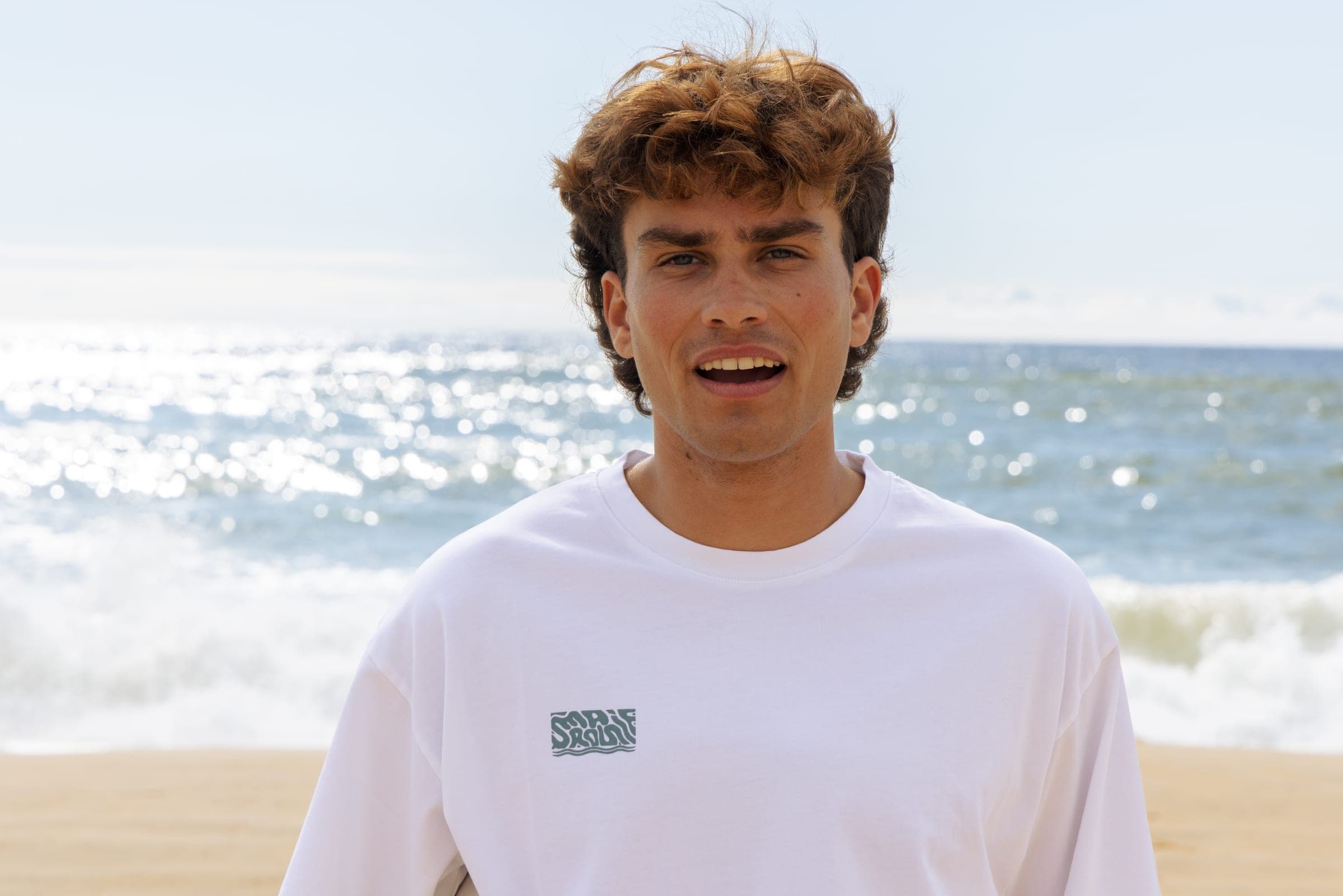 Portrait d'un surfeur en t-shirt blanc, la mer scintille derrière lui.
