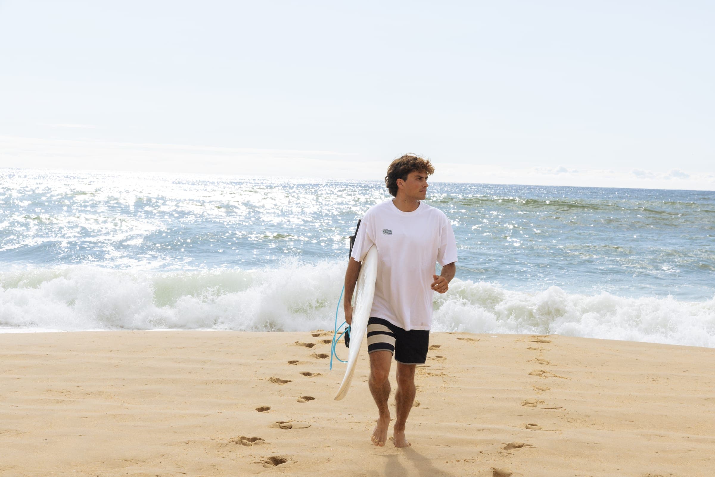 Un surfeur marche vers l'eau, planche à la main, sur le sable mouillé.