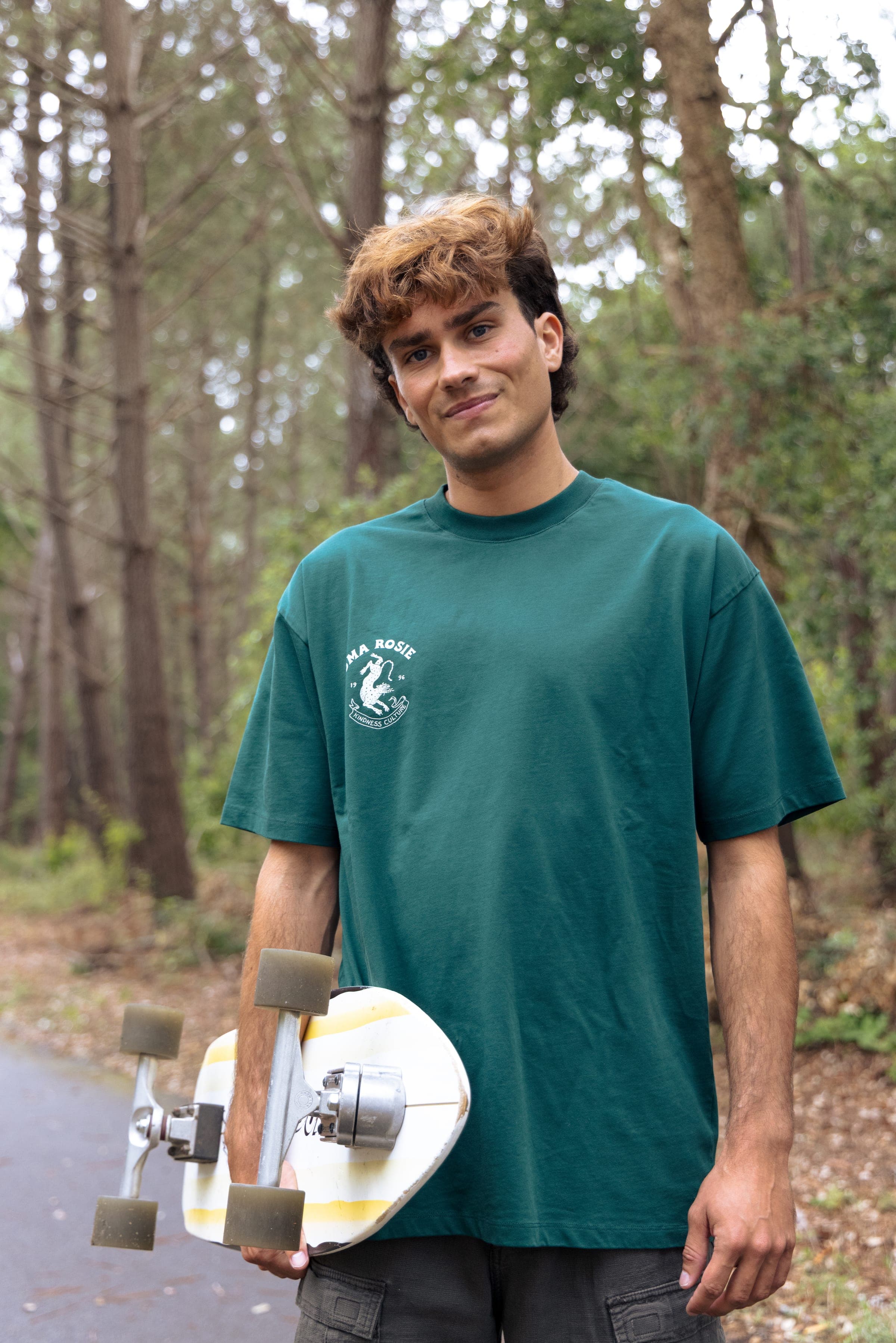 Un skateur en t-shirt vert, longboard sous le bras, dans la forêt de pins.