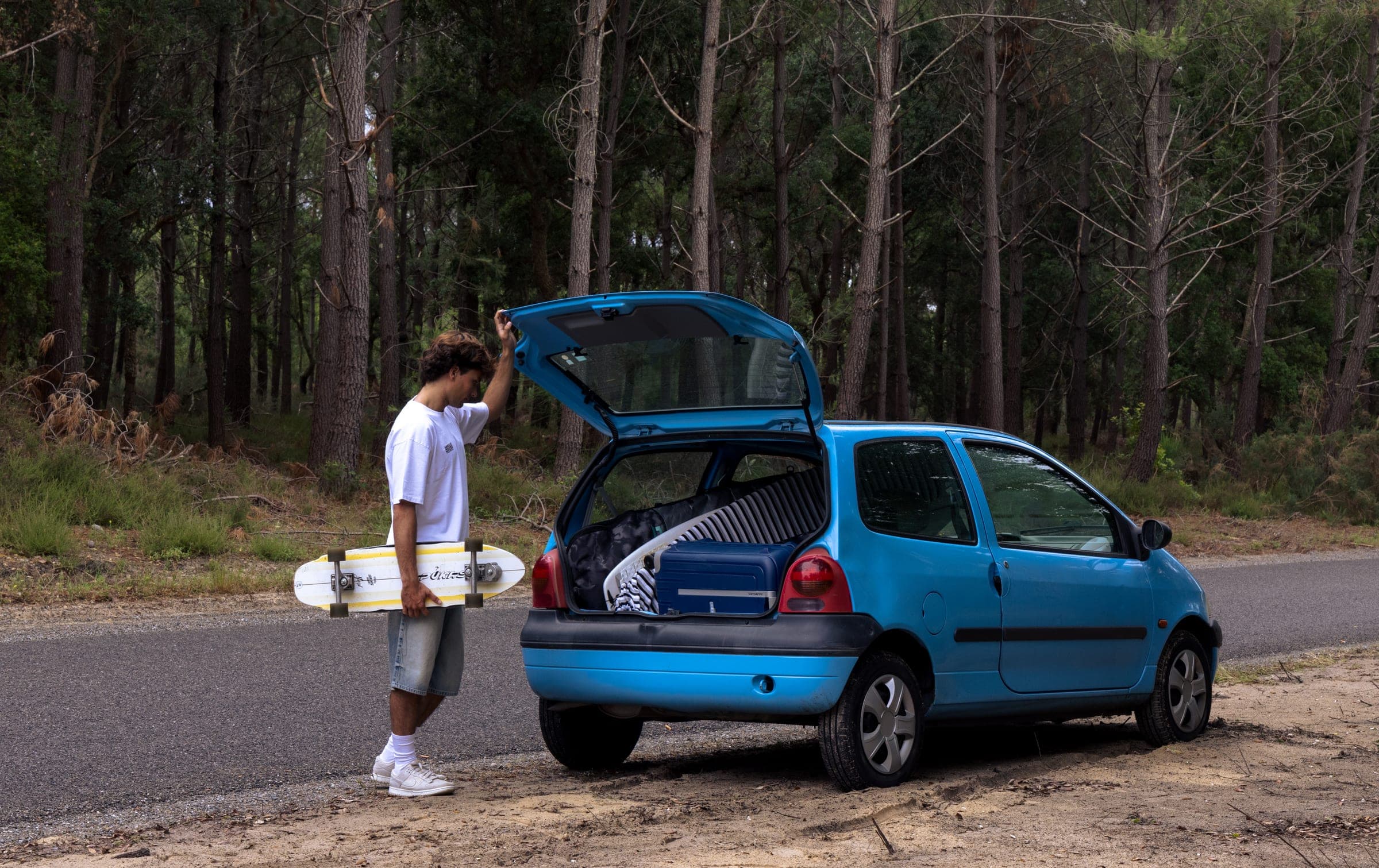 Coffre ouvert d'une Twingo bleue au bord de la route, une planche à la main.