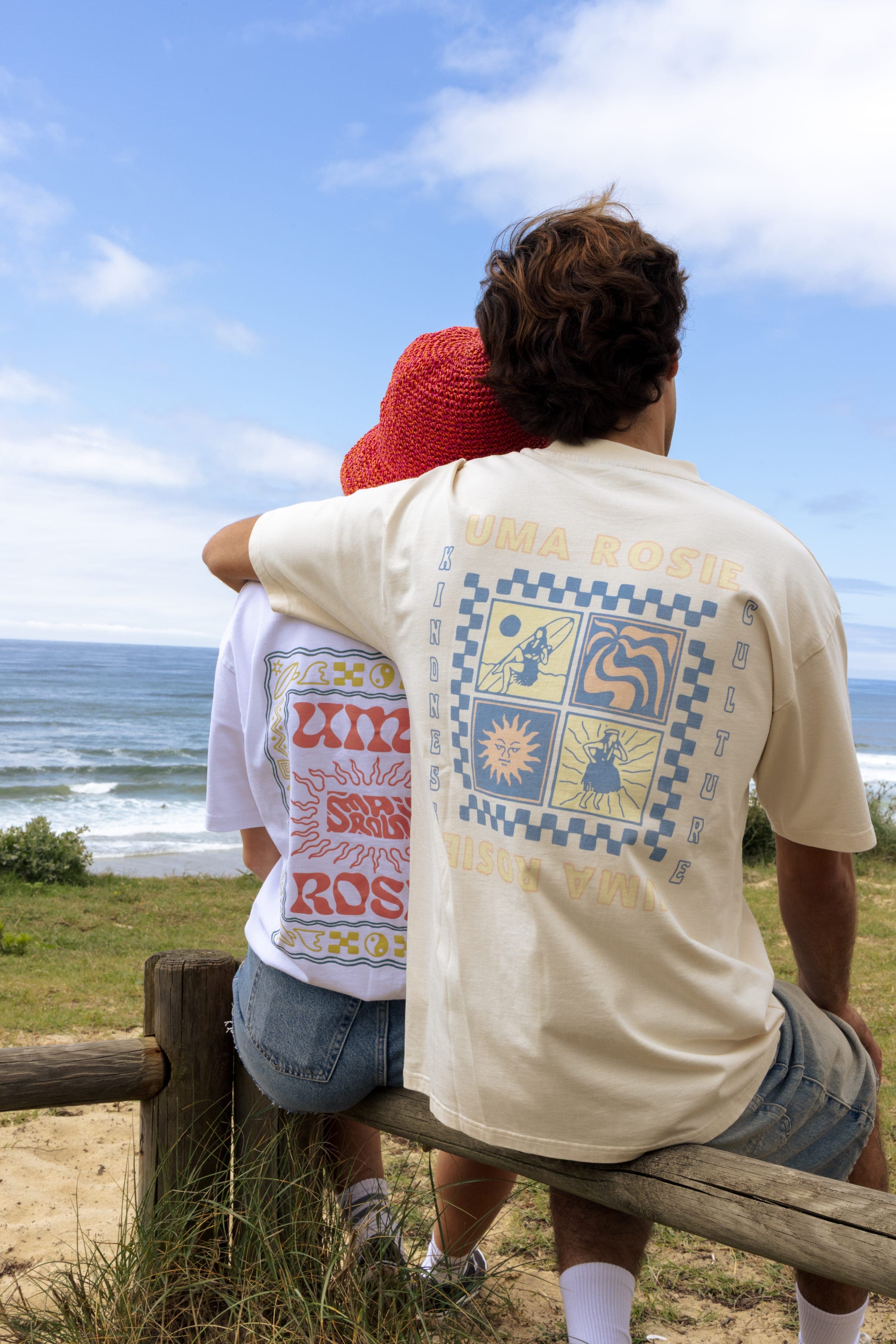 Un couple assis sur une barrière face à l'océan, t-shirt écru sérigraphié de dos.
