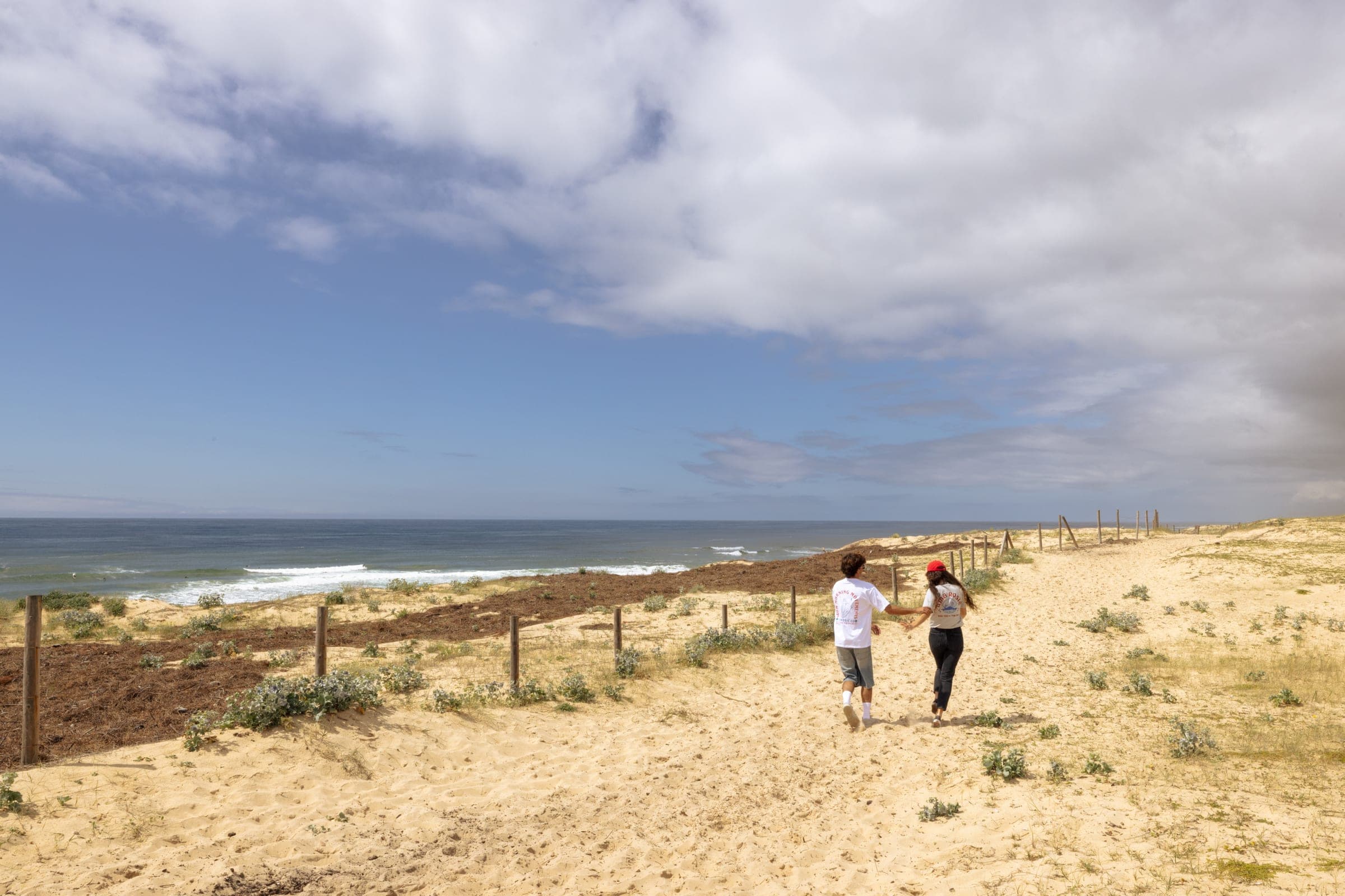 Deux silhouettes marchent vers la plage entre les dunes.