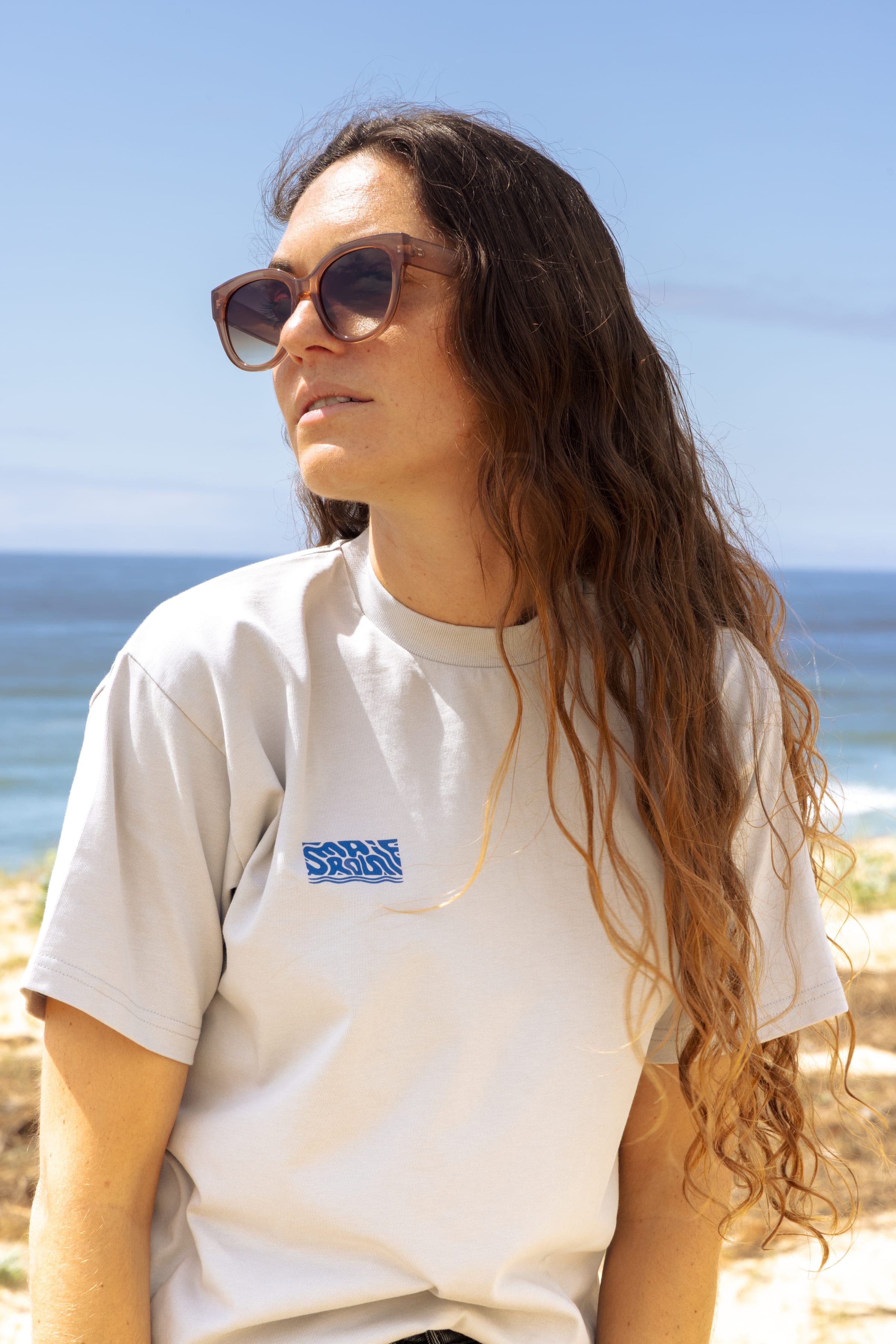 Portrait d'une femme en t-shirt écru et lunettes de soleil, cheveux au vent.