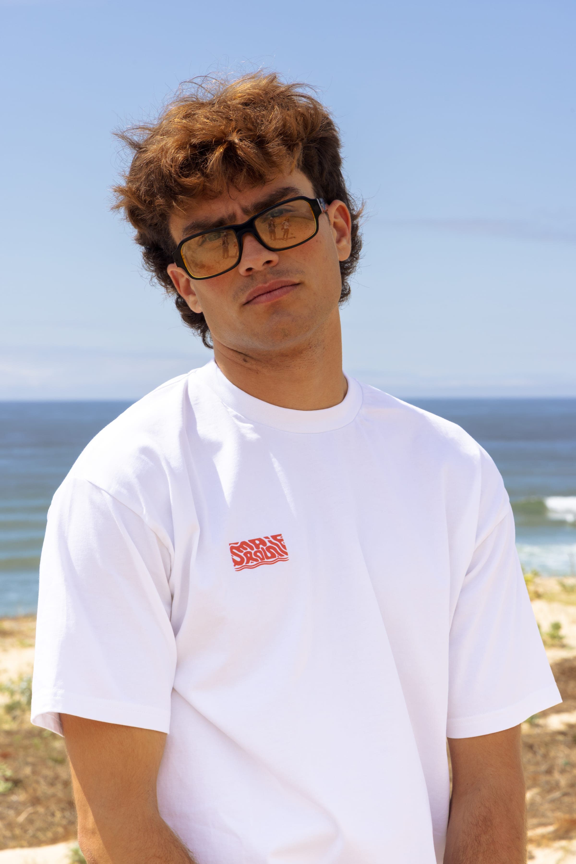 Portrait d'un homme à lunettes en t-shirt blanc, la plage derrière lui.