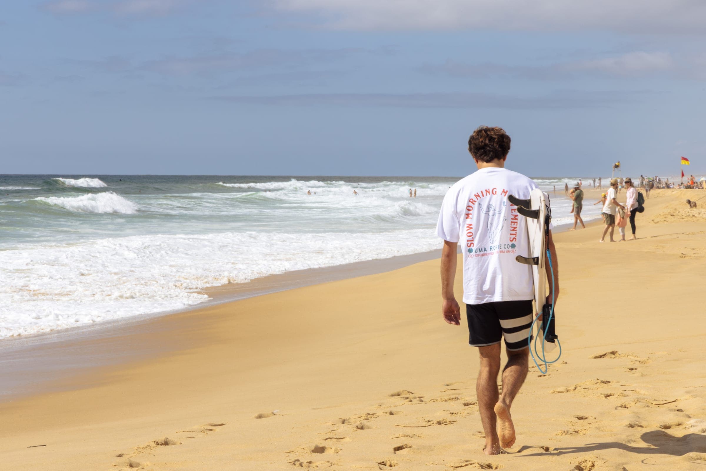 Un surfeur remonte la plage, planche sous le bras, au milieu des baigneurs.