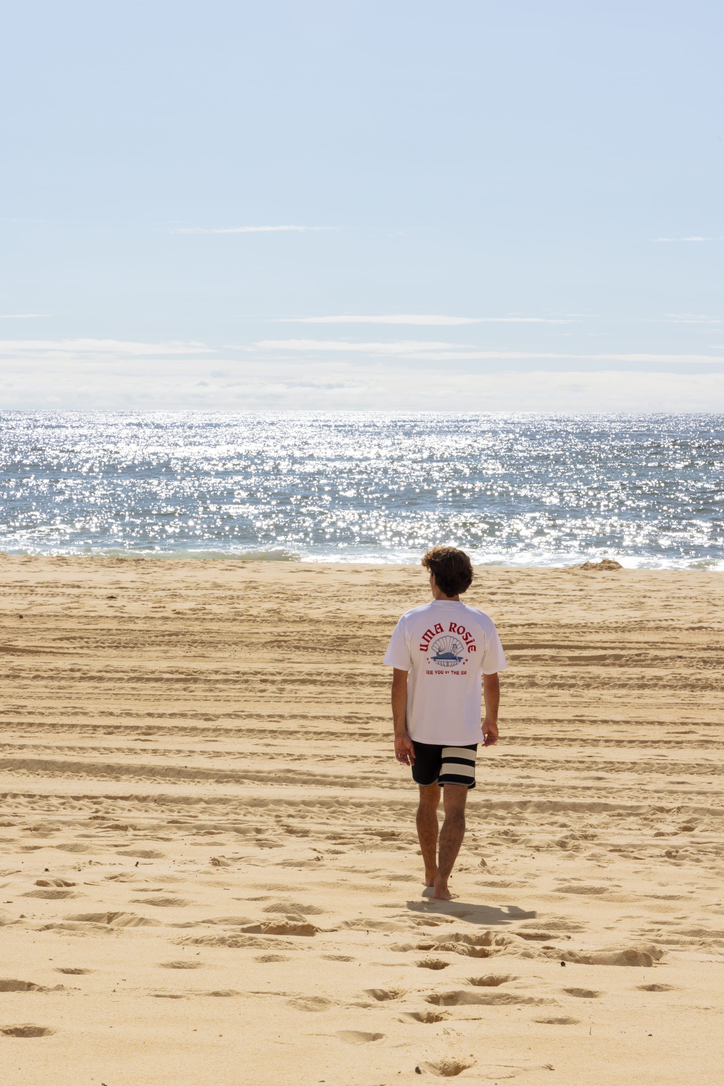 De dos, un surfeur marche sur le sable strié, t-shirt blanc sérigraphié.