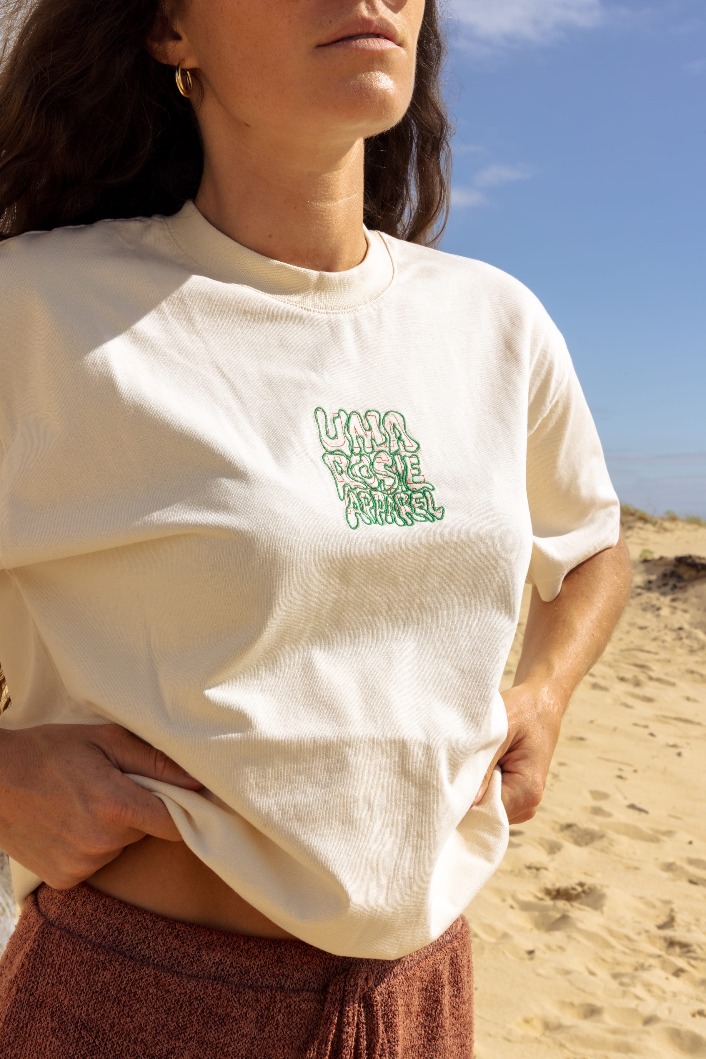 Détail du logo vert brodé sur un t-shirt écru, mains dans le dos.
