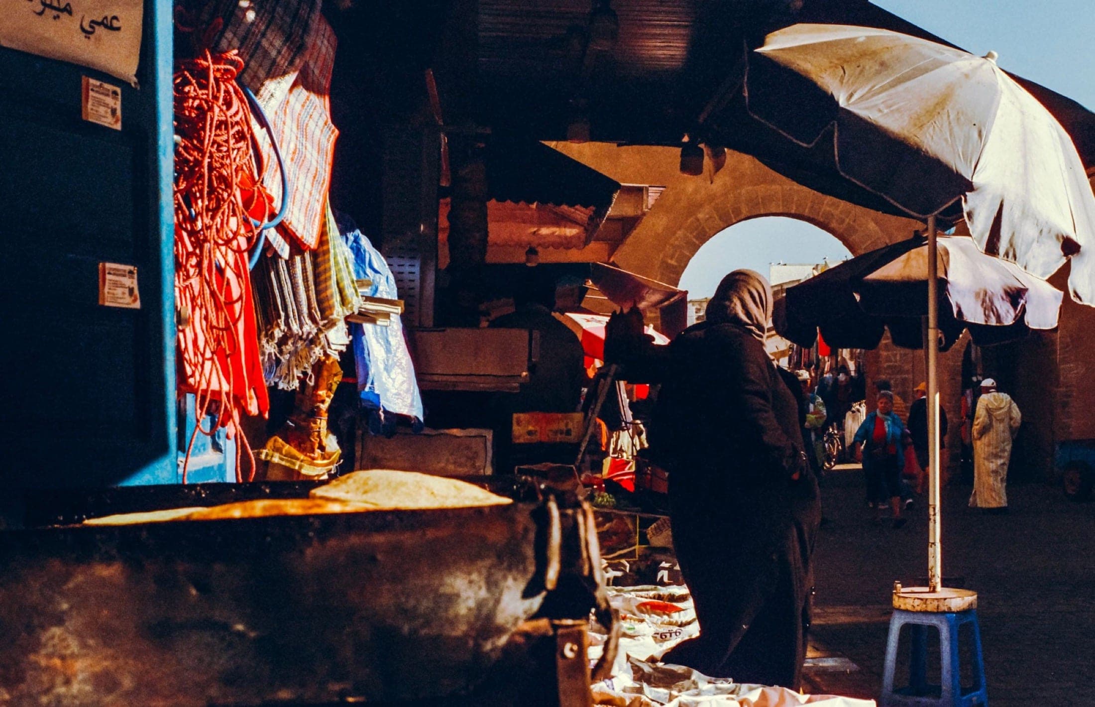 Ruelle de souk à contre-jour, cordes et étoffes suspendues, silhouette de dos sous les parasols, arche de pierre au fond.