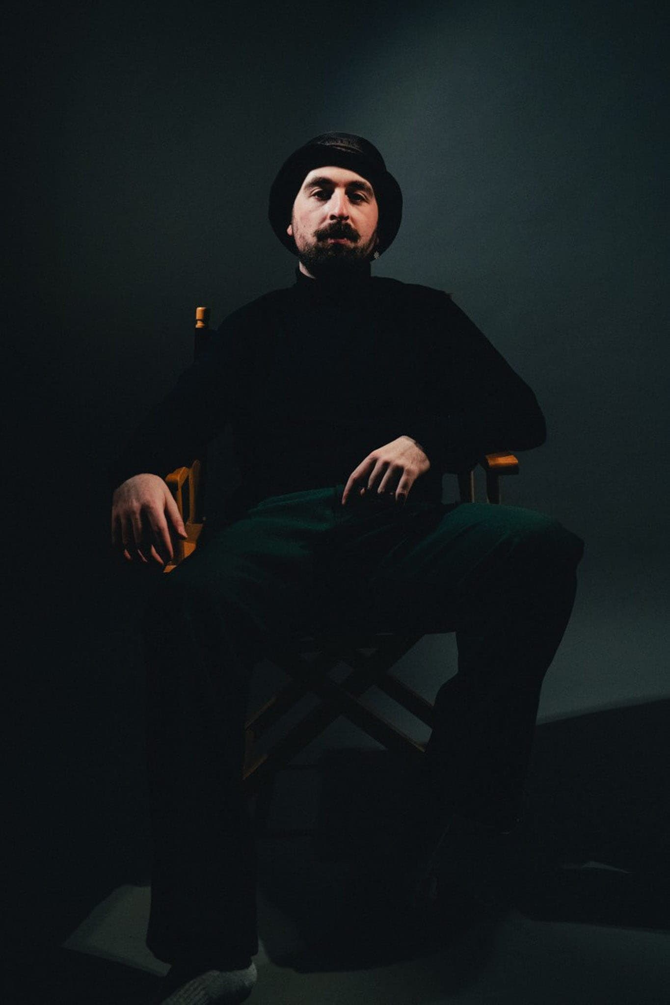 Ruben Haccoun, photographe, assis sur une chaise de réalisateur dans un studio sombre, en bonnet et col roulé noirs, le visage éclairé de côté.