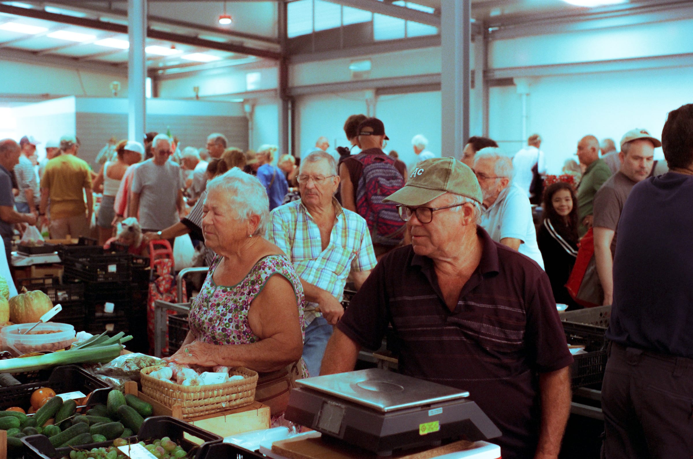 Au marché couvert, une vendeuse et deux clients âgés attendent devant la balance, des concombres sur l'étal.