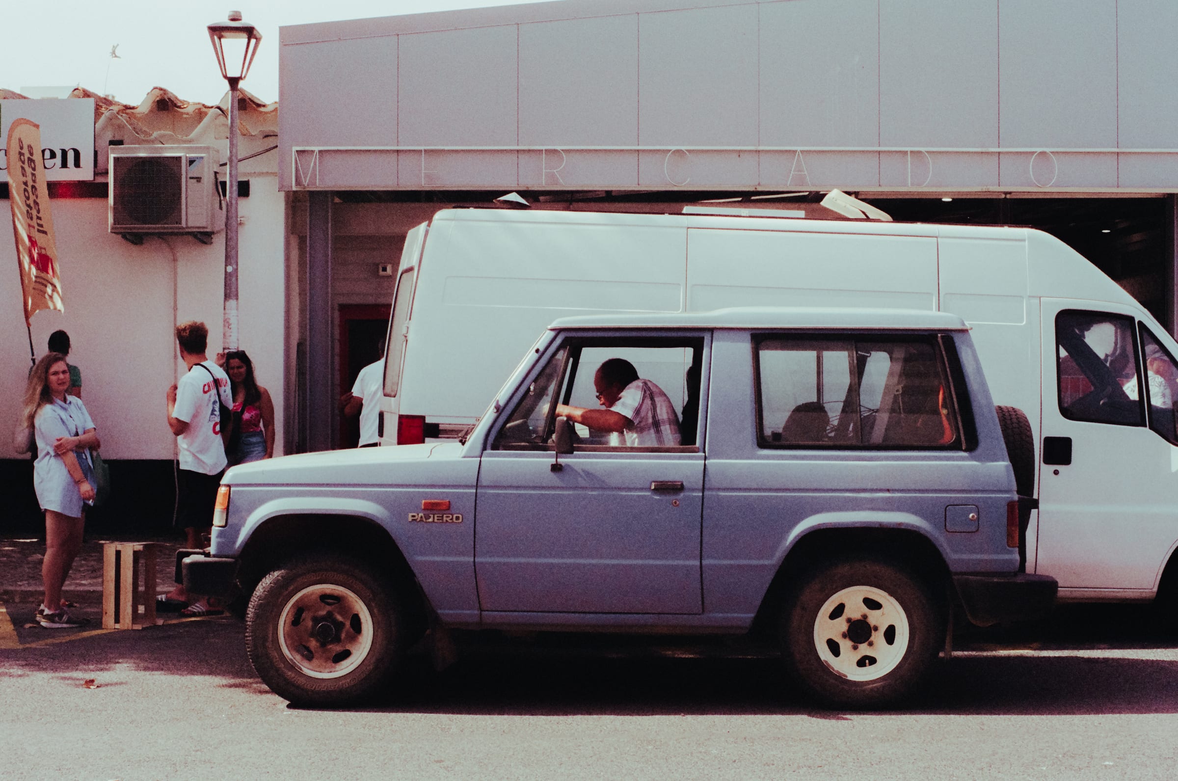 Un vieux Pajero bleu ciel garé devant le Mercado, le conducteur penché vers la portière.