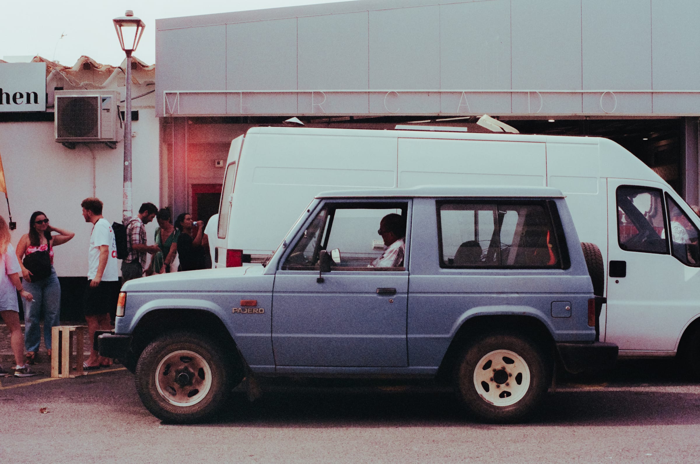 Un Pajero bleu devant l'entrée du marché, des clients discutent sur le trottoir derrière lui.