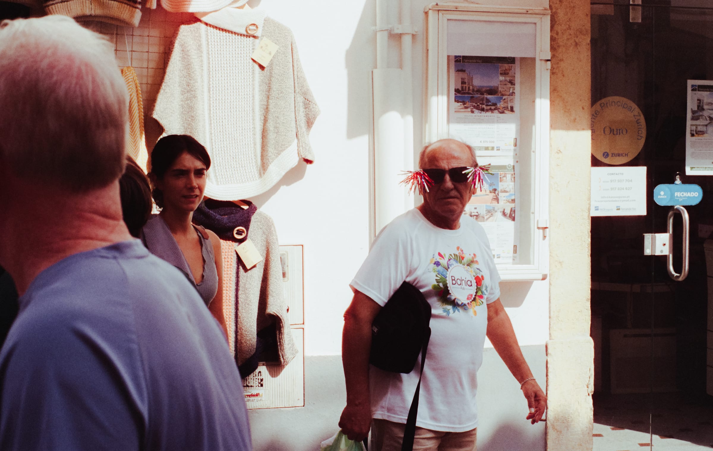 Dans une rue commerçante, un homme en t-shirt imprimé porte des lunettes de soleil hérissées de franges roses.