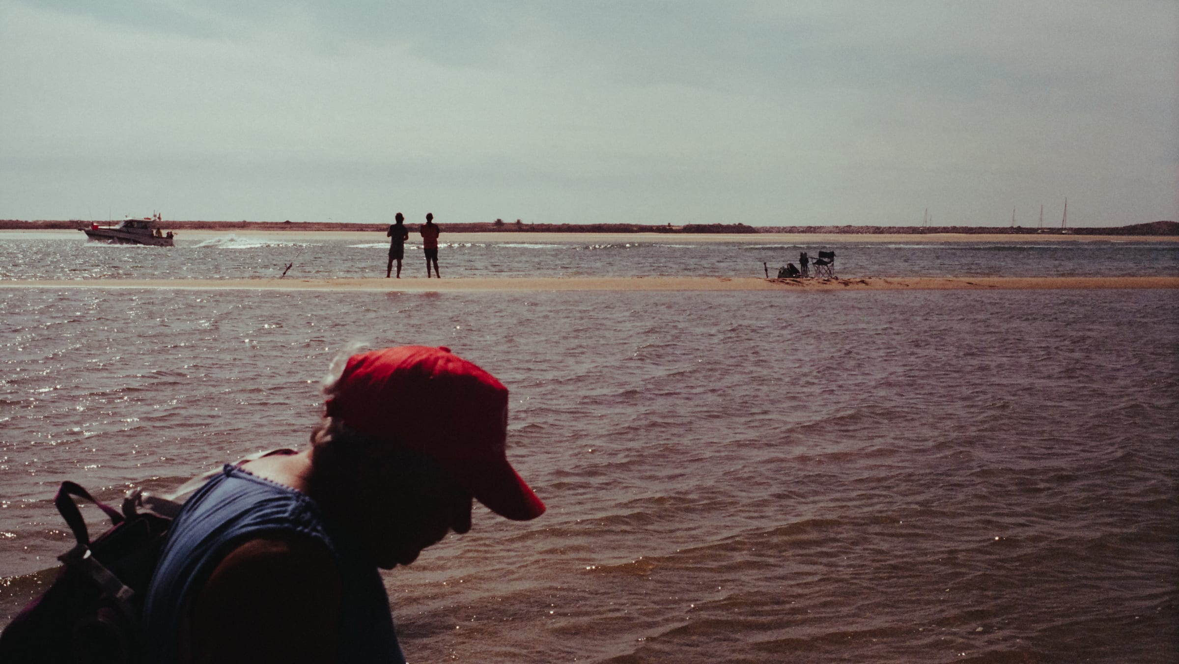 De dos au bord de l'eau, un homme à casquette rouge, deux silhouettes plantées sur le banc de sable au loin.