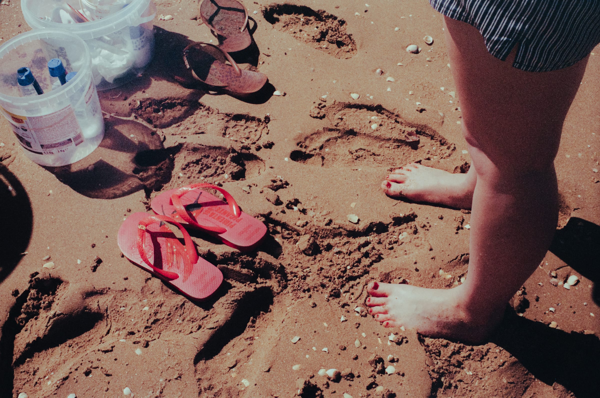 Des tongs roses, des seaux et des pieds nus dans un sable couvert de traces.