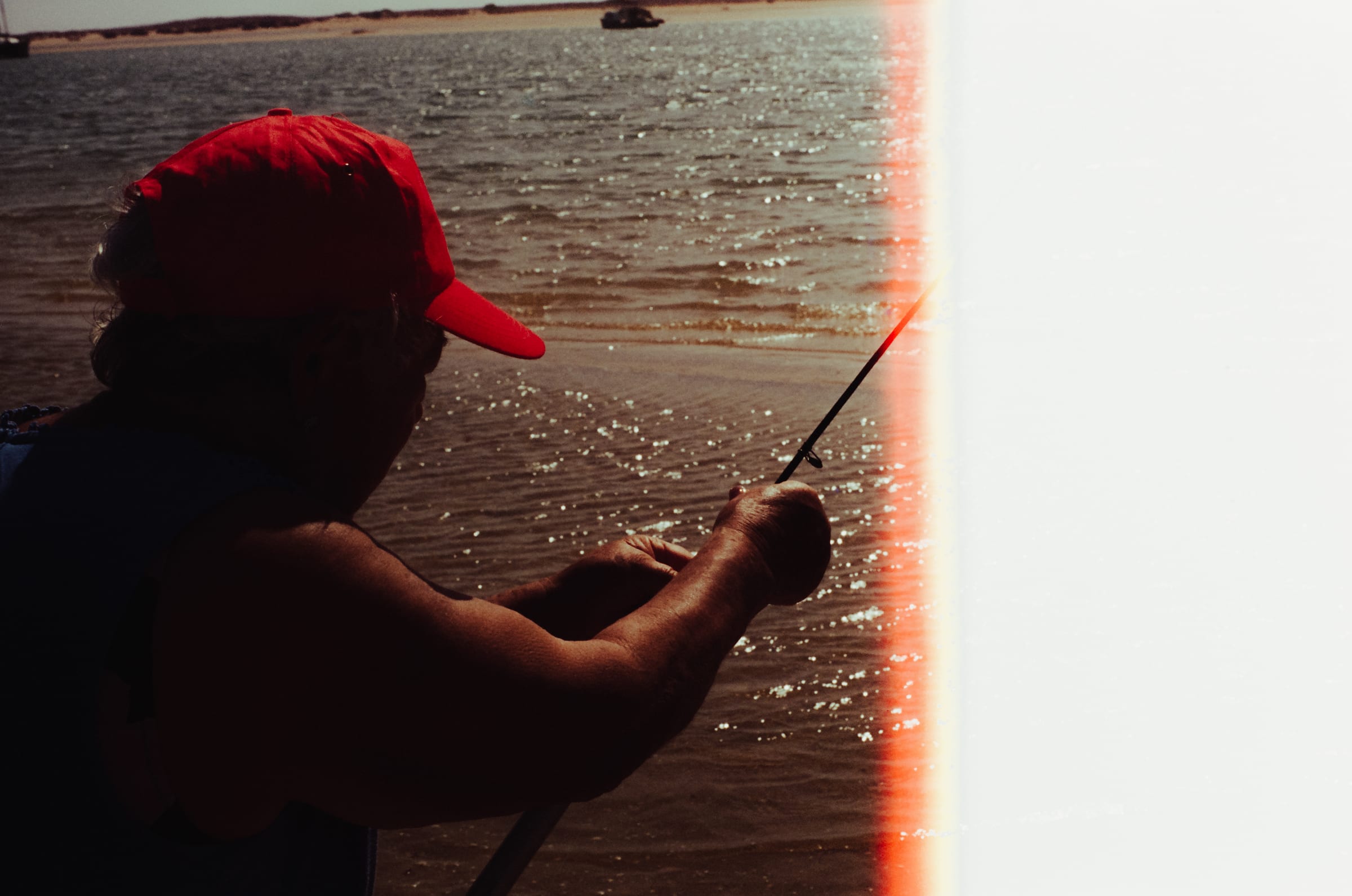 Une silhouette à casquette rouge tend sa canne vers l'eau, la moitié droite de l'image voilée de lumière.