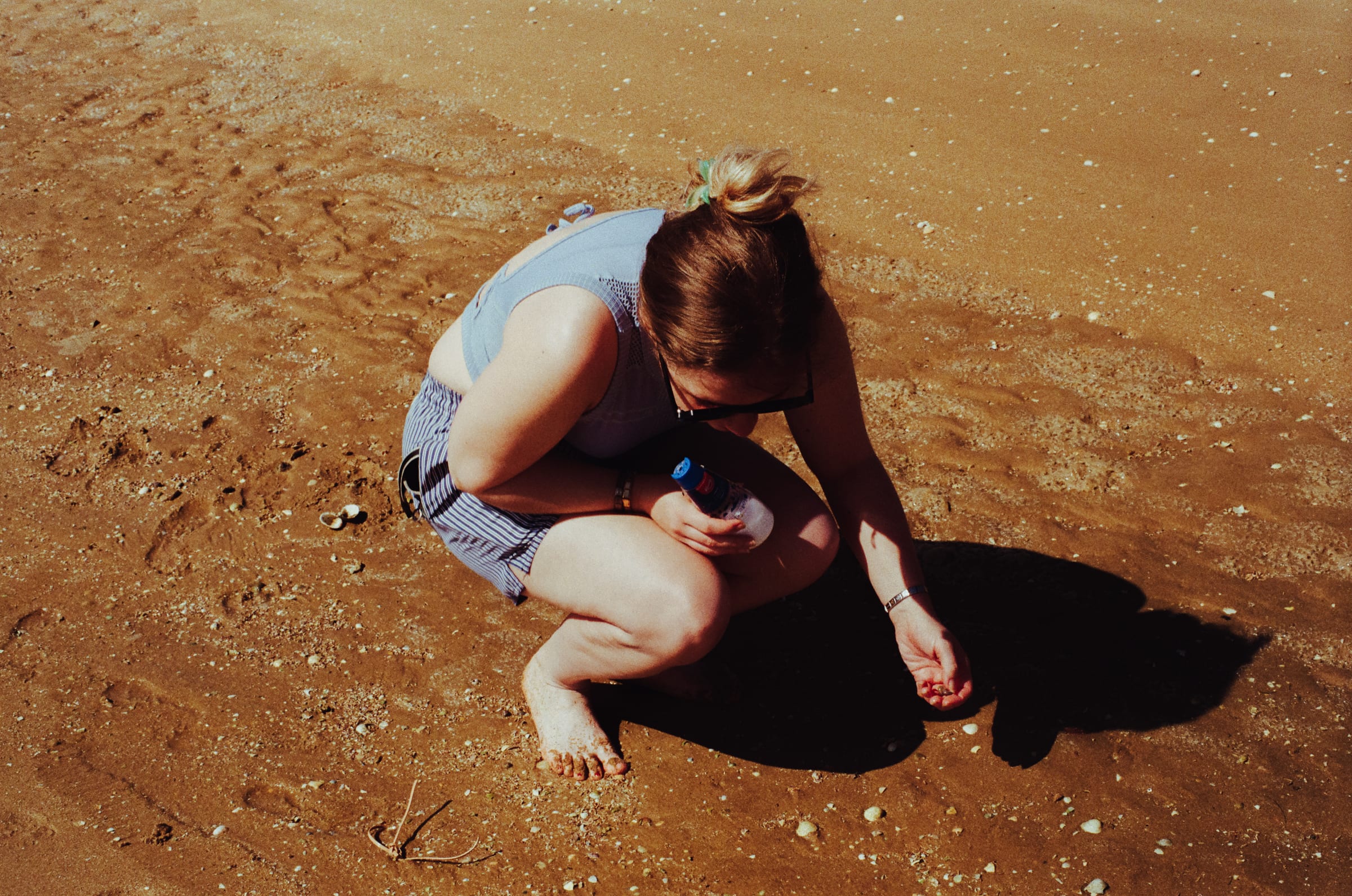 Accroupie sur le sable mouillé, une jeune femme cherche des couteaux, un flacon à la main.