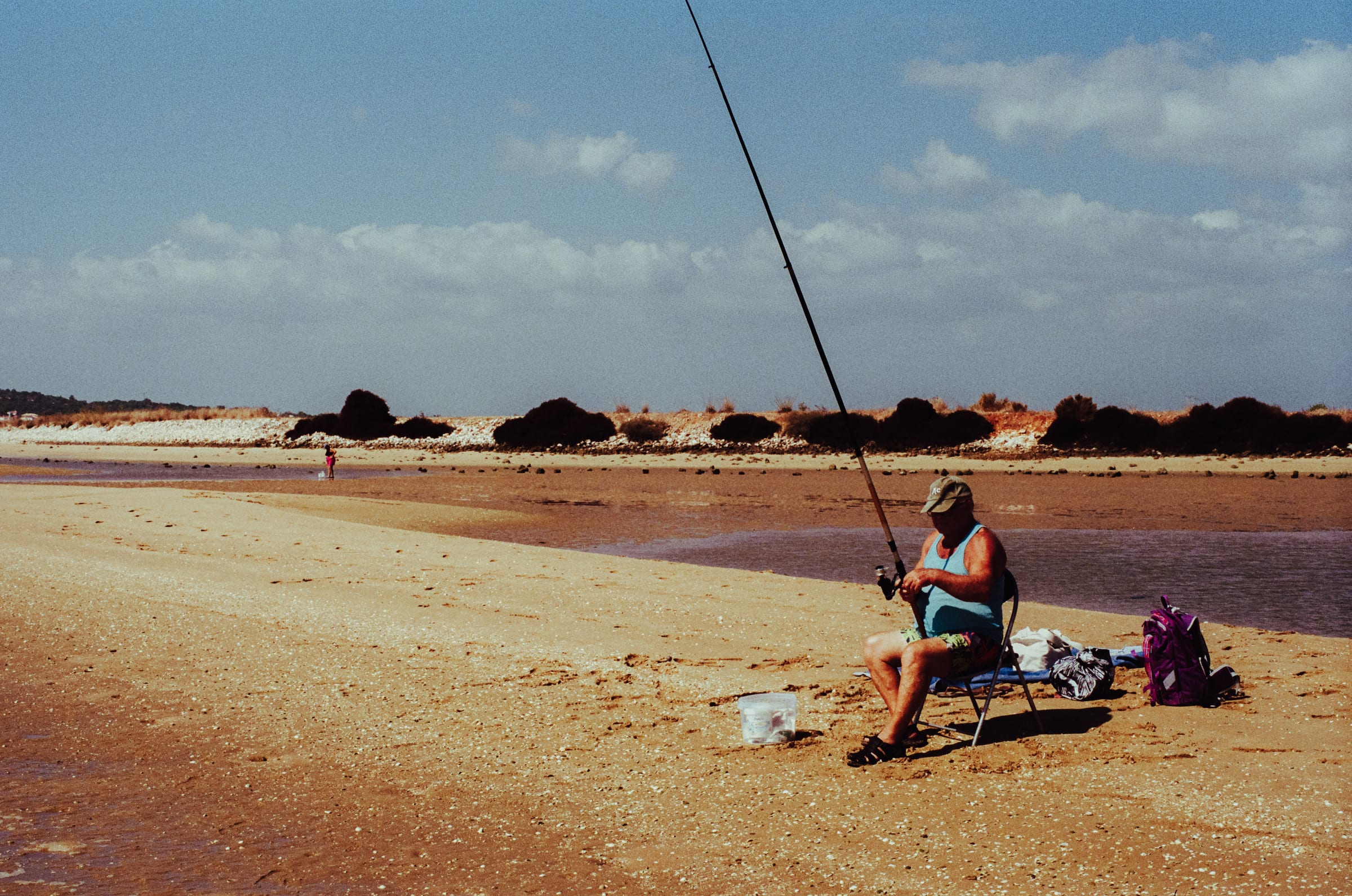 Un pêcheur sur sa chaise pliante, canne dressée vers le ciel, sur le banc de sable à marée basse.