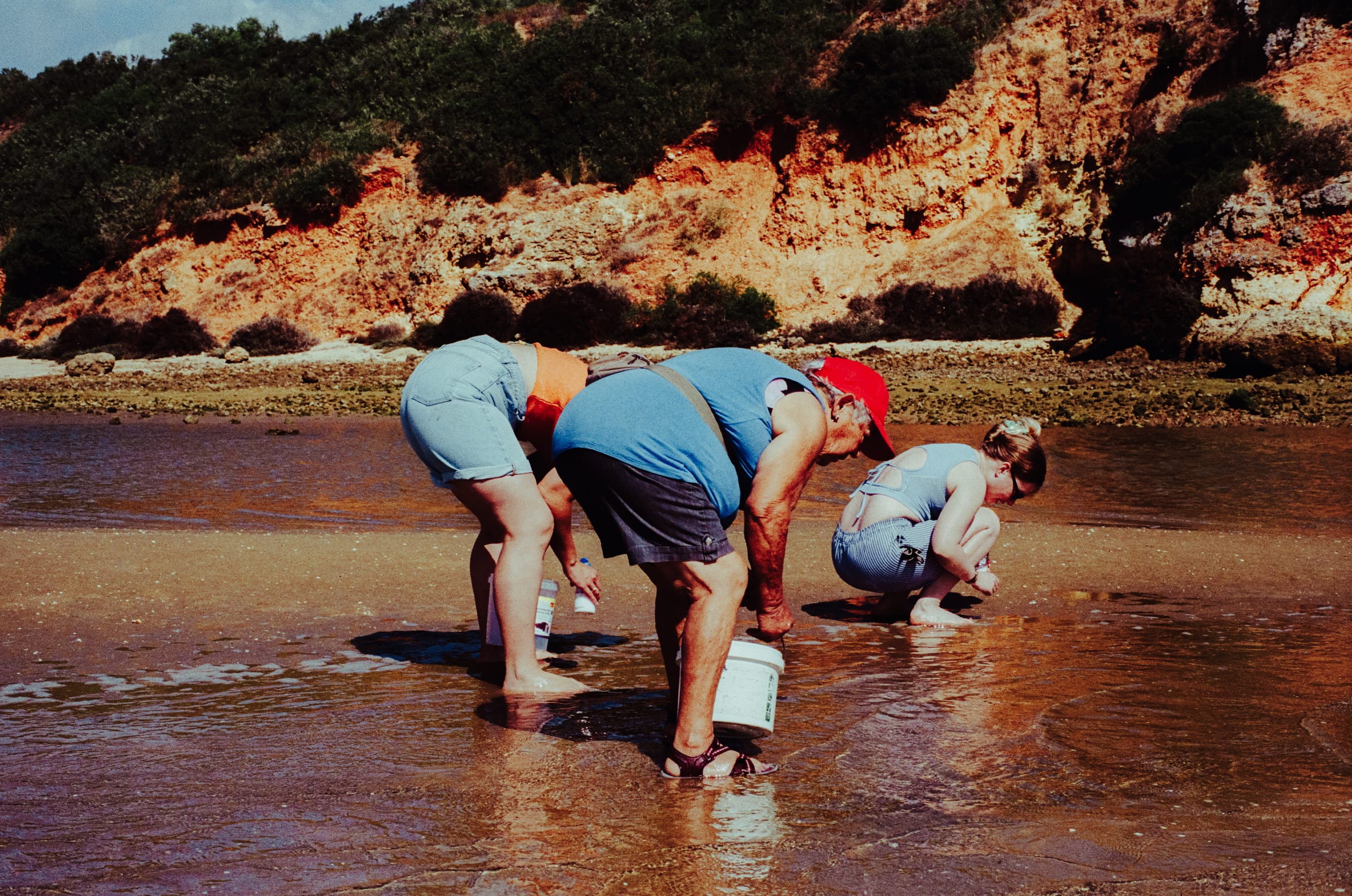 Trois personnes courbées sur le sable mouillé au pied d'une falaise ocre, à la pêche aux couteaux.