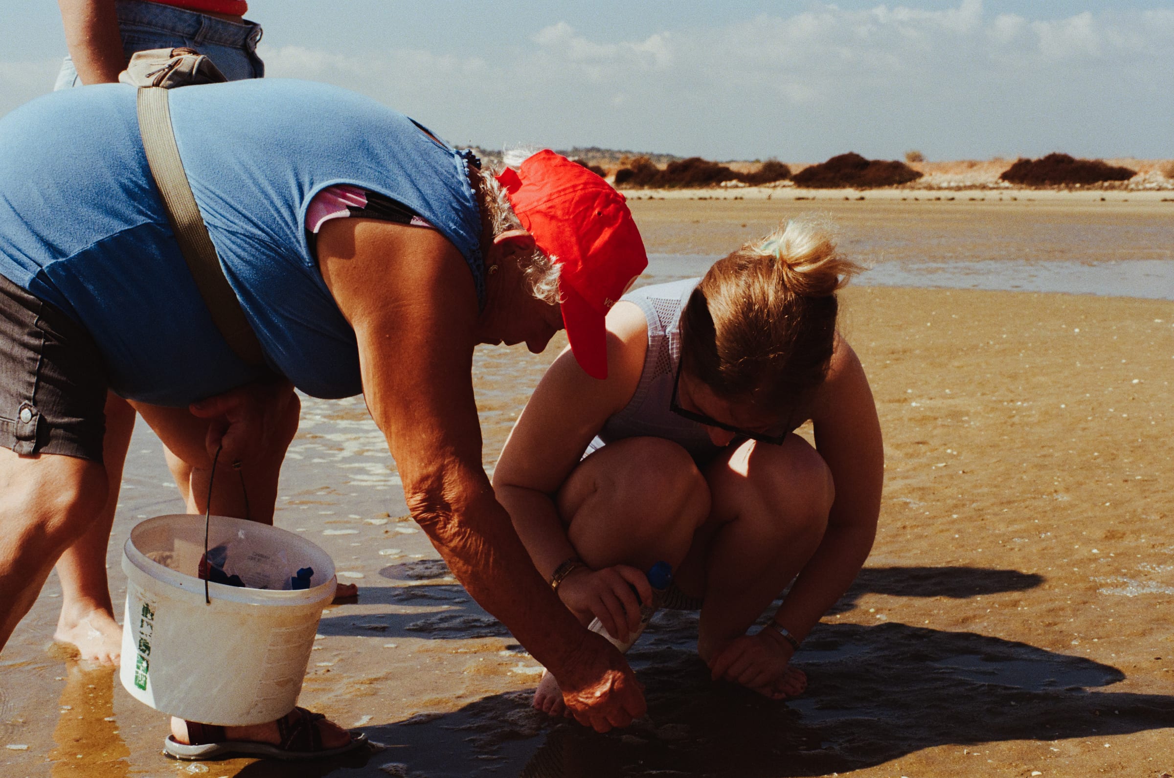 Une femme à casquette rouge et une jeune femme accroupie fouillent le sable, un seau entre elles.