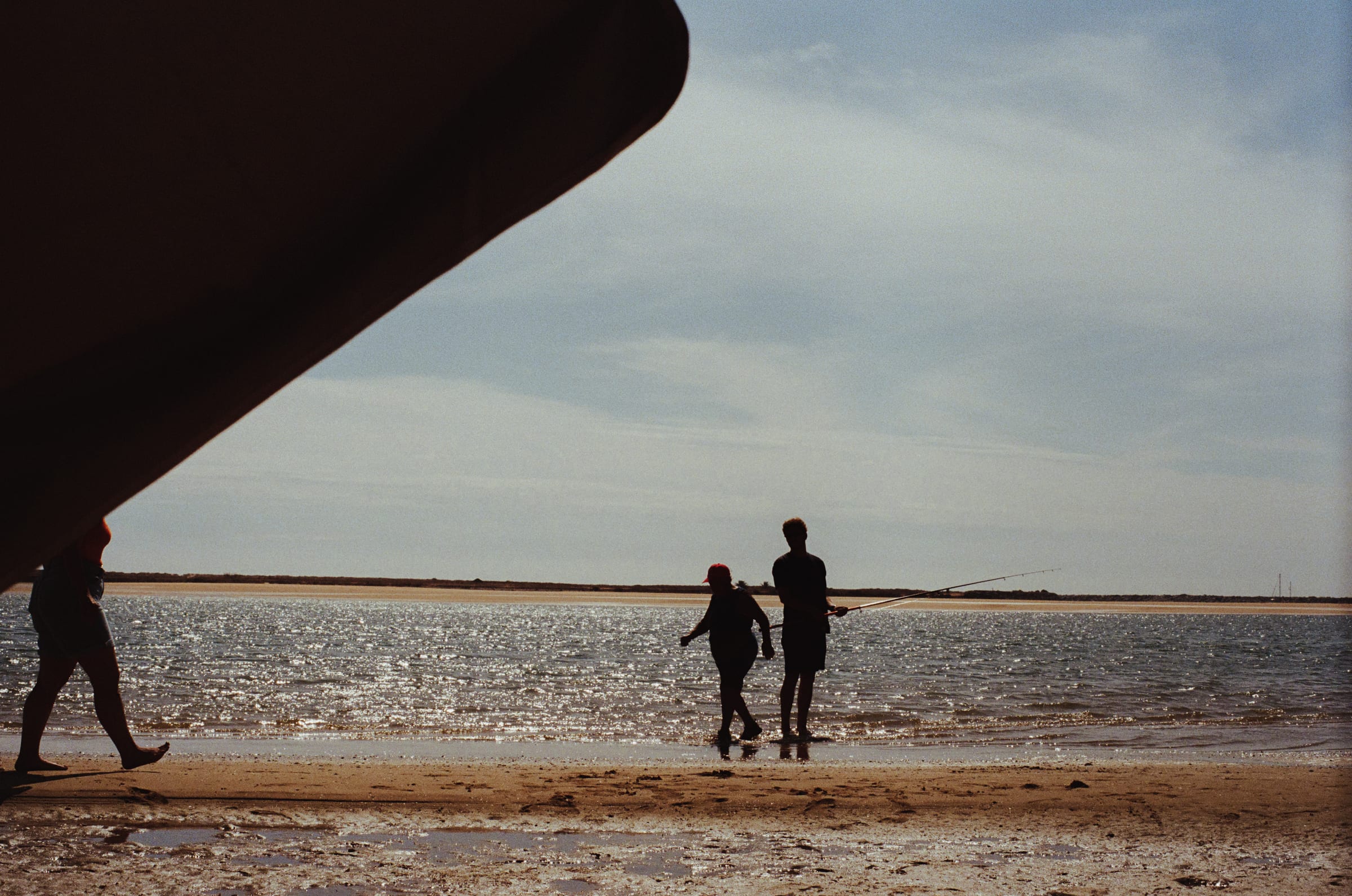 Sous le bord d'un parasol, deux pêcheurs en silhouette au bord de l'eau pendant qu'un baigneur passe.