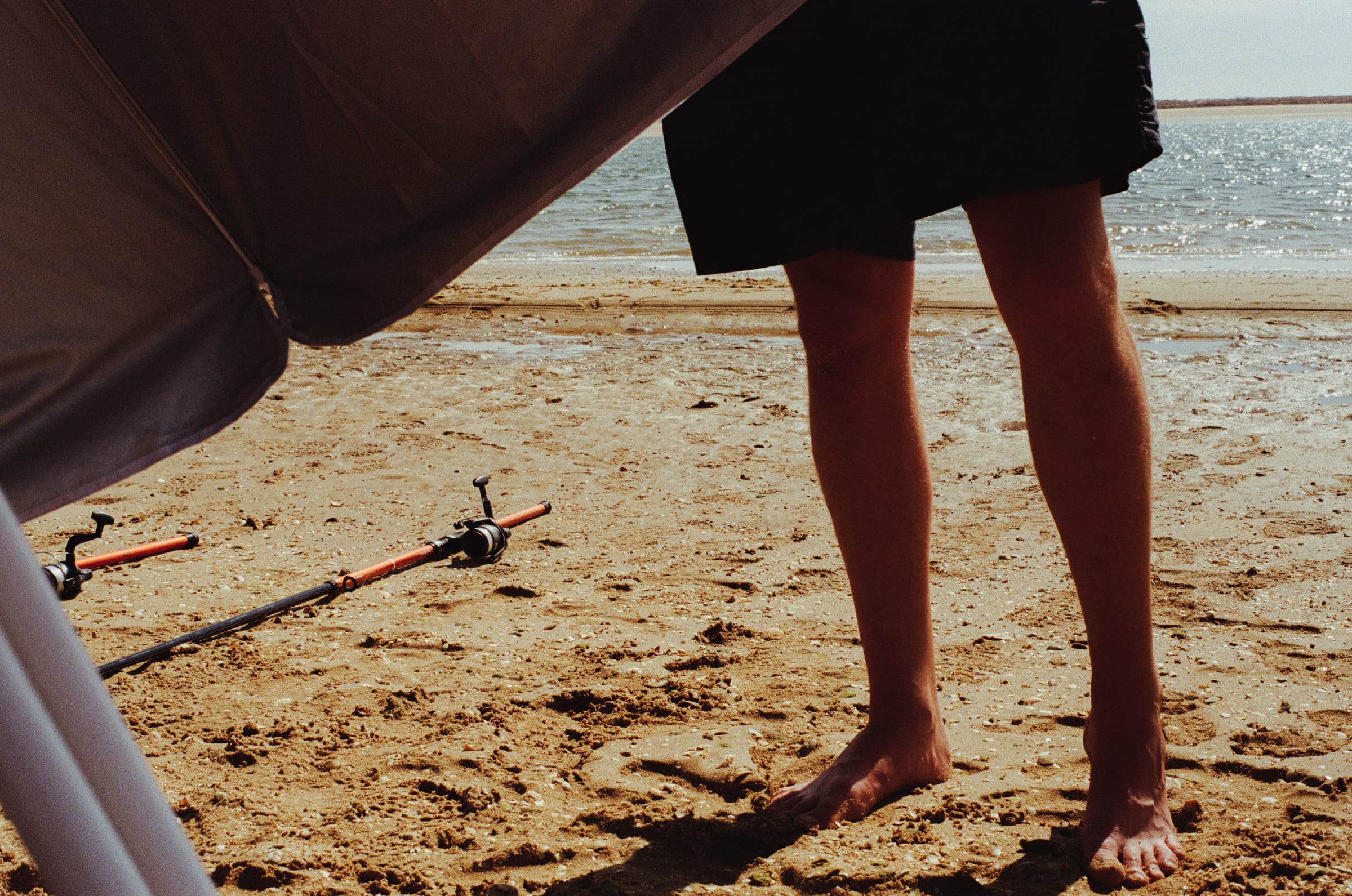 Des jambes nues à l'ombre d'un parasol, deux cannes à pêche posées dans le sable.